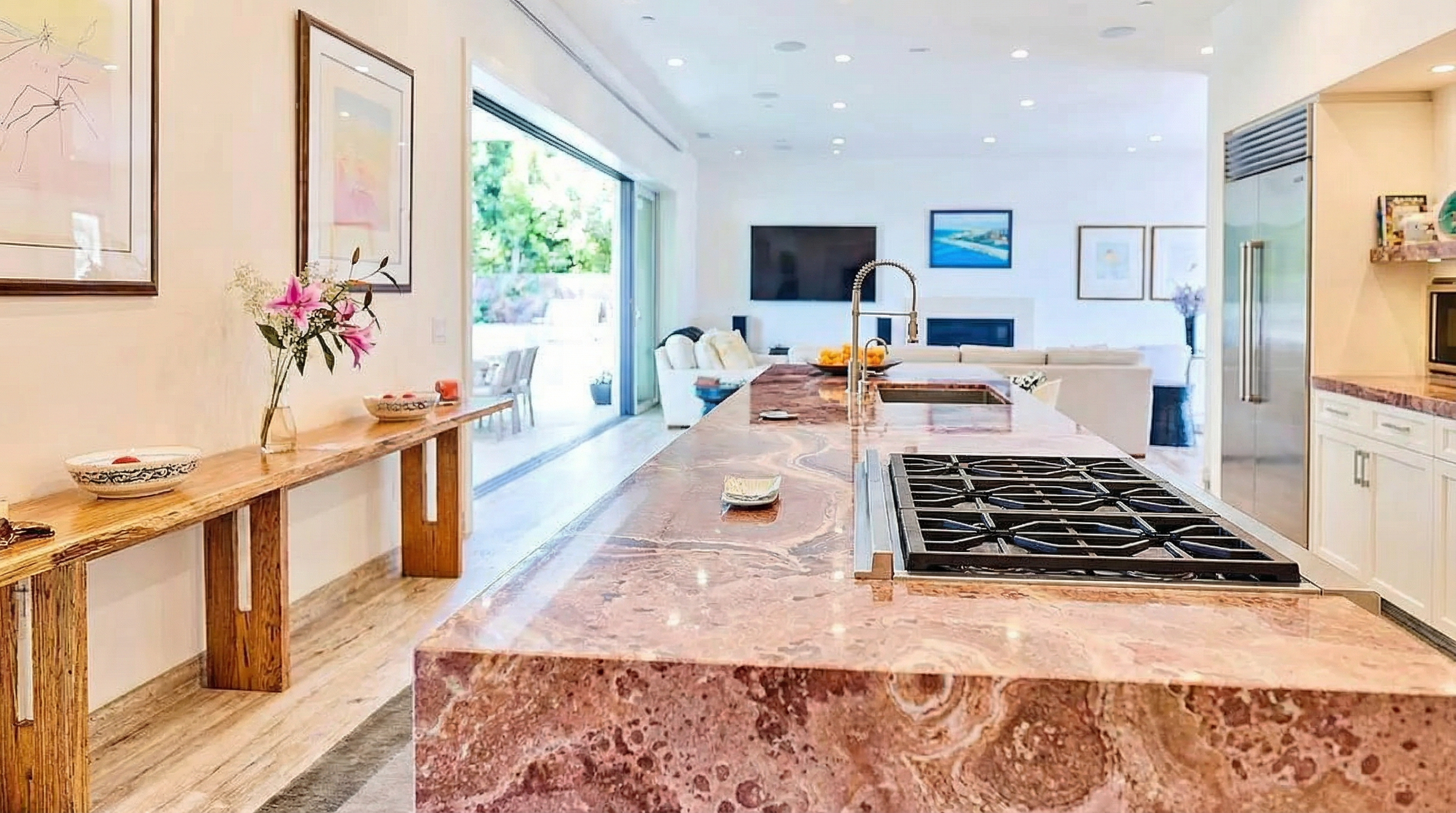 This is a bright, contemporary kitchen featuring a large island with a unique reddish-brown marble countertop. Modern appliances are present, and the kitchen opens to a living area with a sliding glass door leading to an outdoor space. The kitchen's design emphasizes a spacious and open floor plan, ideal for entertaining and family living.