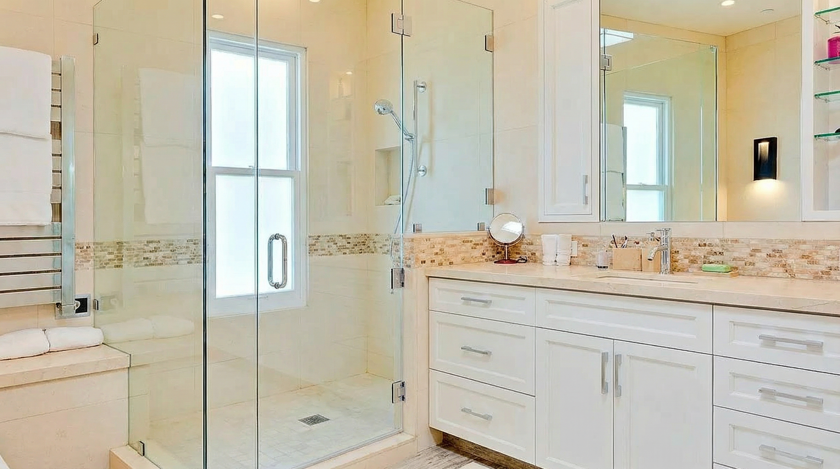 This is a well-lit primary bathroom featuring a walk-in shower with glass enclosure and a double vanity with white cabinets. The countertop is a light color, and there's a decorative tile backsplash. The overall design is modern and clean, creating a spa-like atmosphere.