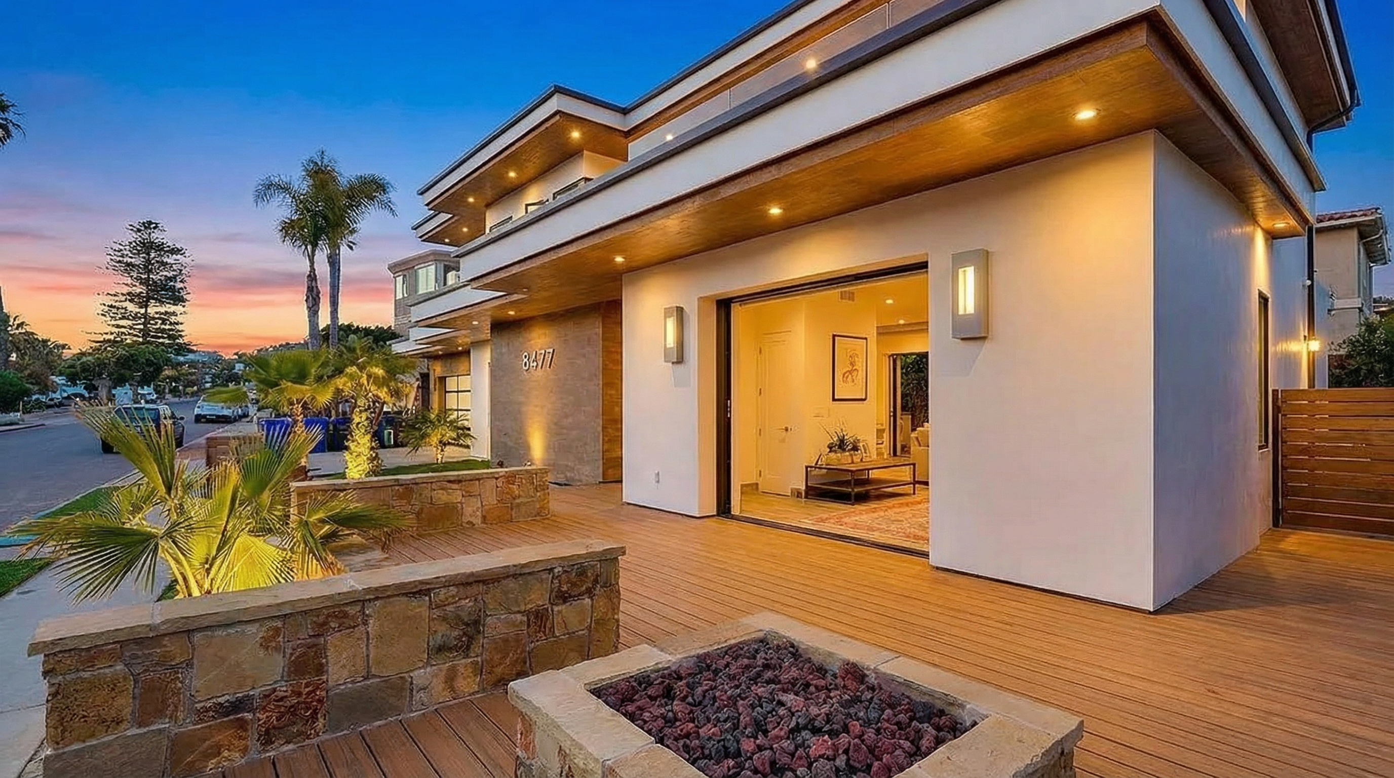 This image showcases the front exterior of a modern residence. The architectural design highlights clean lines, a combination of white walls and wood accents, and a multi-level structure. The outdoor space features a deck with a built-in fire pit and landscaping, contributing to a welcoming and stylish curb appeal.