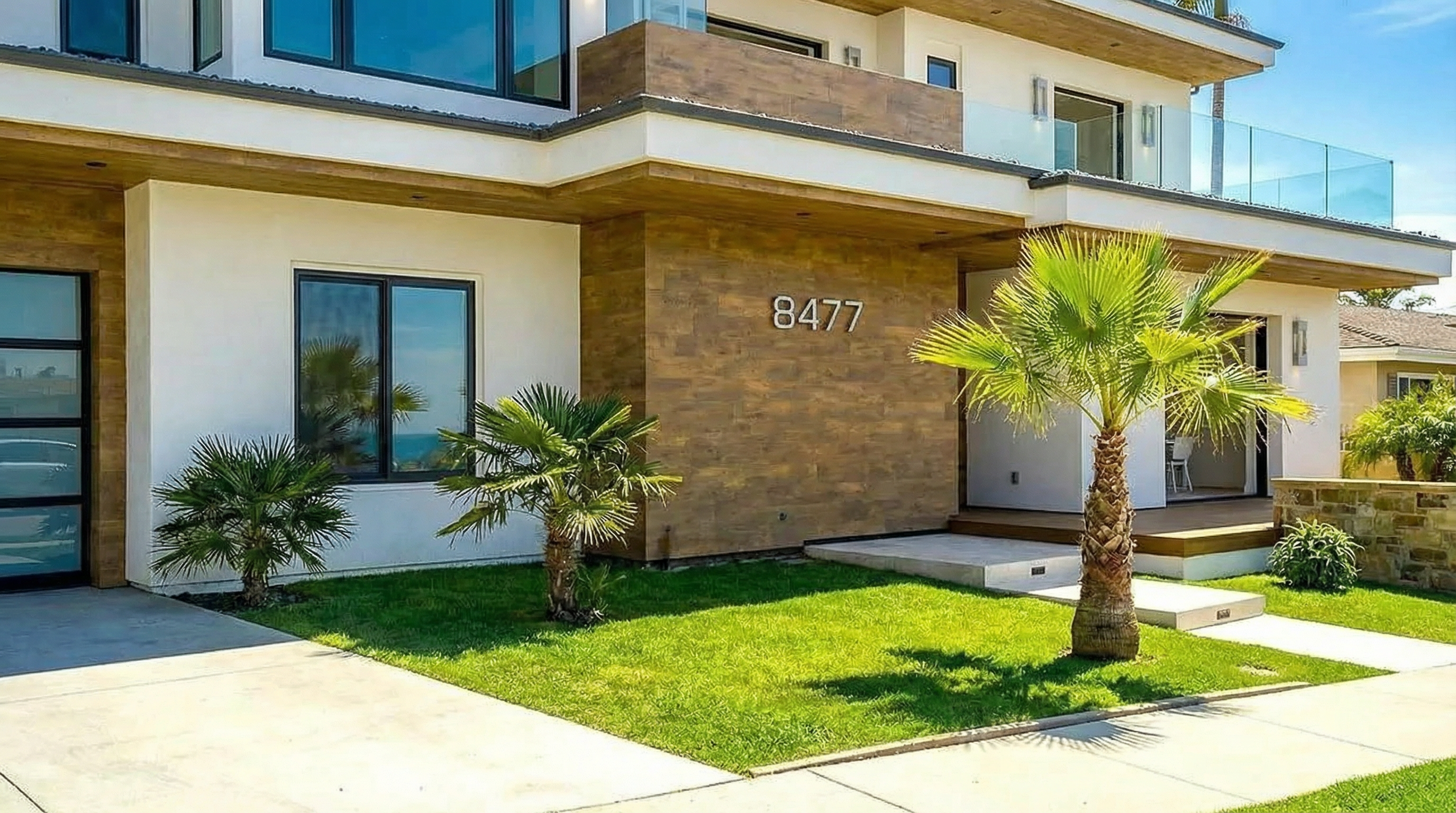 This is the front exterior of a modern, two-story house. The facade features a combination of white stucco and wood-look siding, with a prominent address number '8477'. Landscaping includes small palm trees and well-maintained green lawn, enhancing the curb appeal.