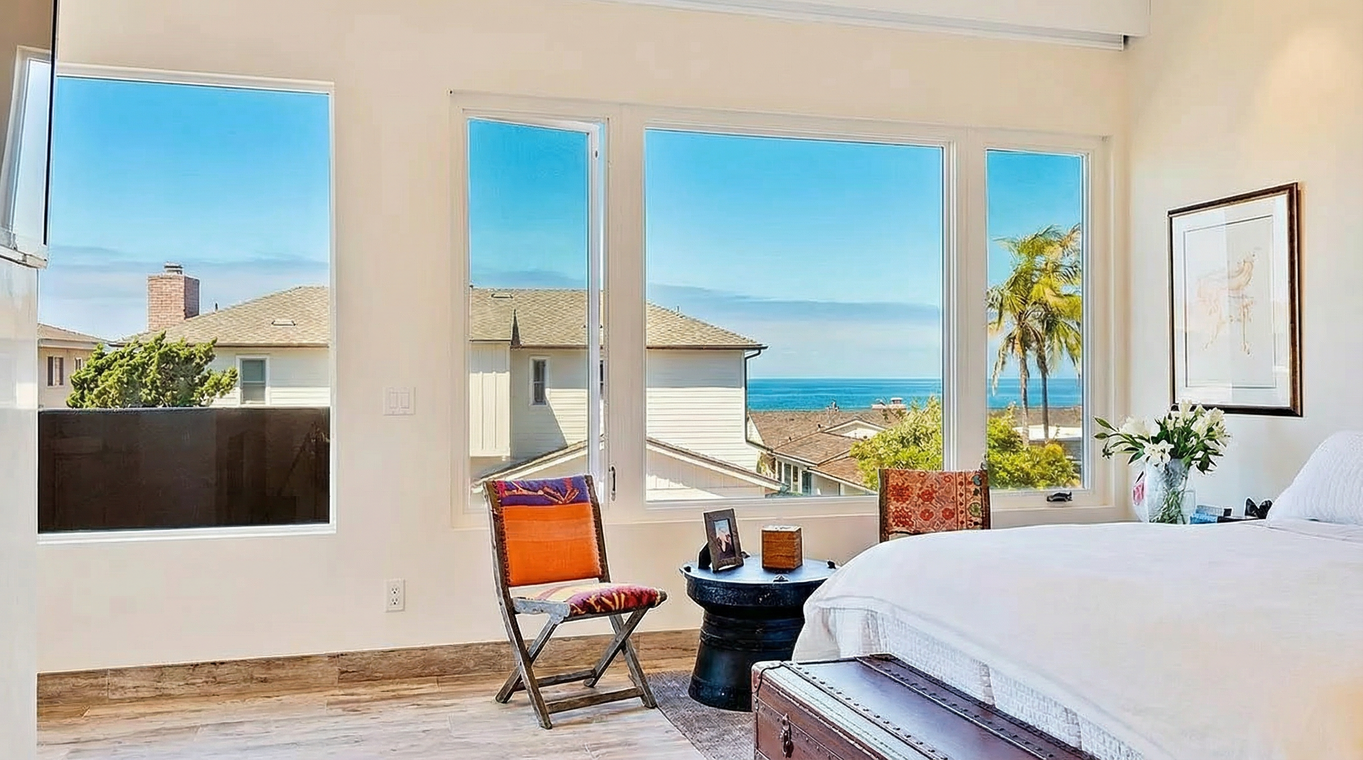 This is a well-lit primary bedroom with a large window offering a scenic view of the ocean and surrounding houses. The room features comfortable furniture, including a bed, chairs, and a unique drum-shaped side table. The hardwood flooring and artwork add a touch of elegance and sophistication to the space.
