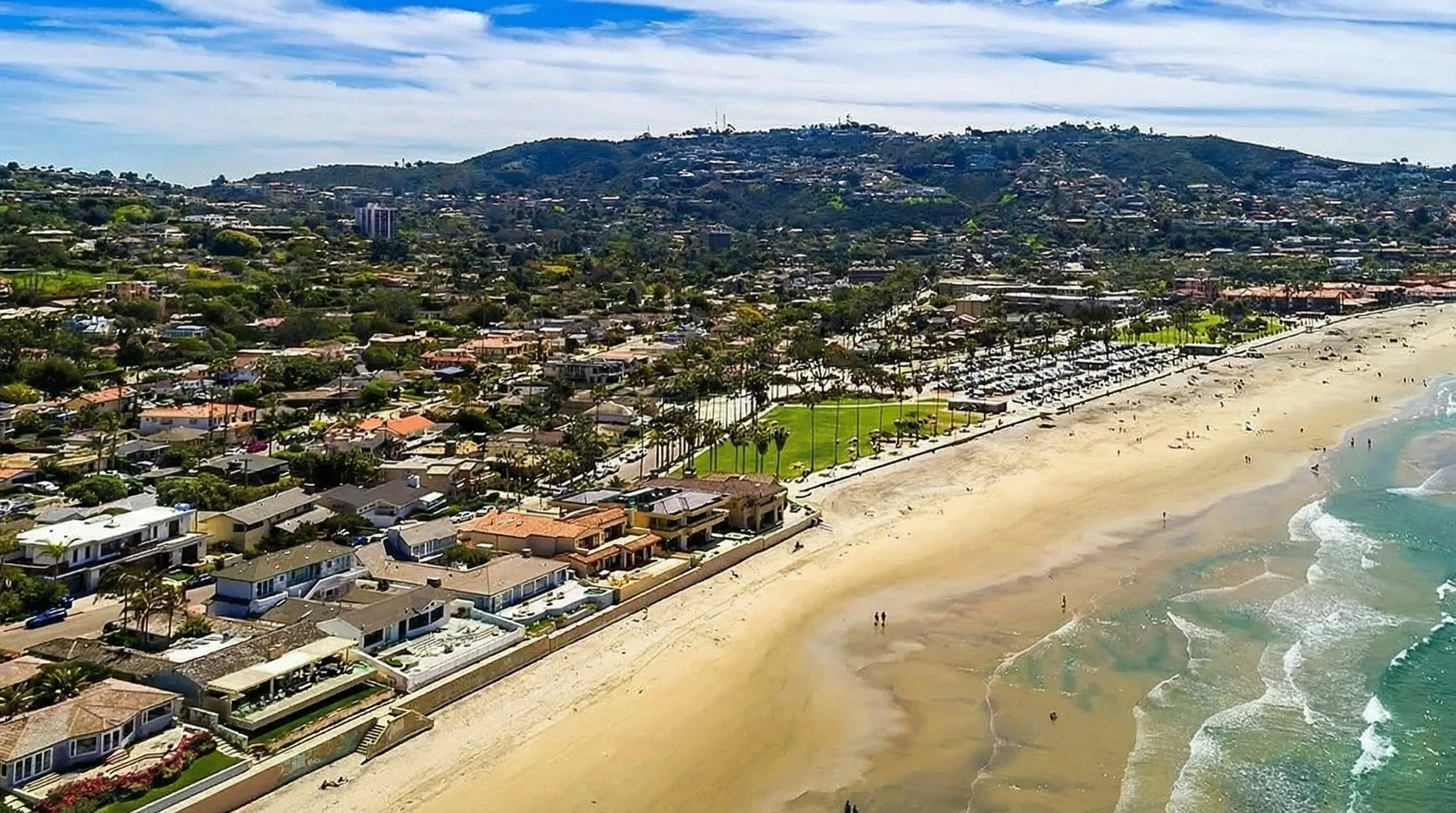 This aerial view captures a stunning coastal property featuring multiple homes along a sandy beach. Palm trees line a grassy area adjacent to a parking lot, adding to the serene seaside atmosphere. The homes appear luxurious, and the beach stretches invitingly with gentle waves meeting the shoreline.