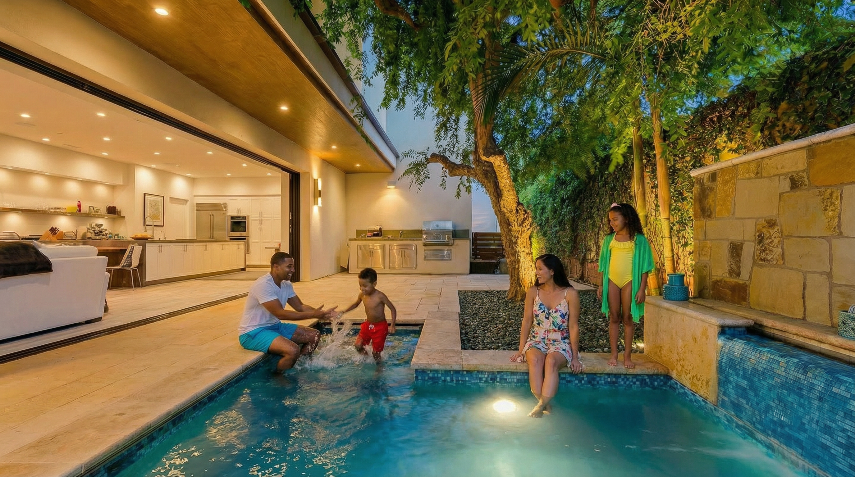 This image showcases a luxurious outdoor pool area, featuring a tiled pool with a waterfall feature. The pool is framed by stone coping and surrounded by lush landscaping, including mature trees. Part of the modern house with a large sliding door opening to the kitchen is visible. The overall impression is one of relaxation and high-end outdoor living.