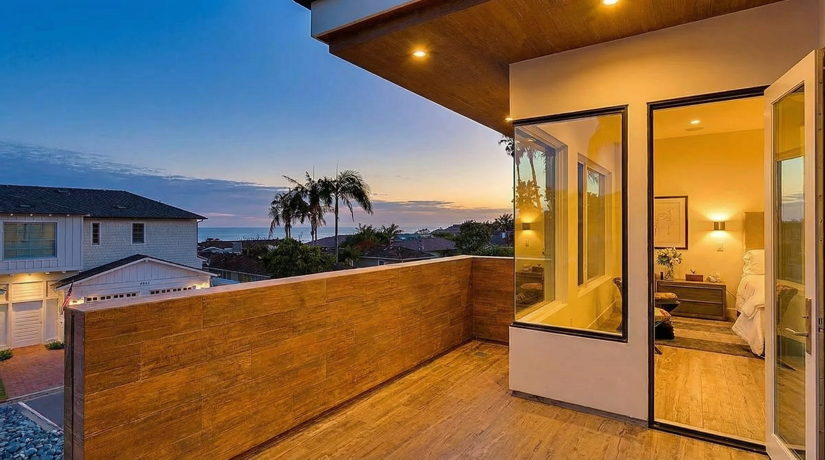This image showcases a beautifully designed balcony with wood-look tiling and a matching privacy wall. The open doors lead into a brightly lit bedroom, seamlessly blending indoor and outdoor space. The view includes residential rooftops, palm trees, and a glimpse of the ocean providing natural beauty.