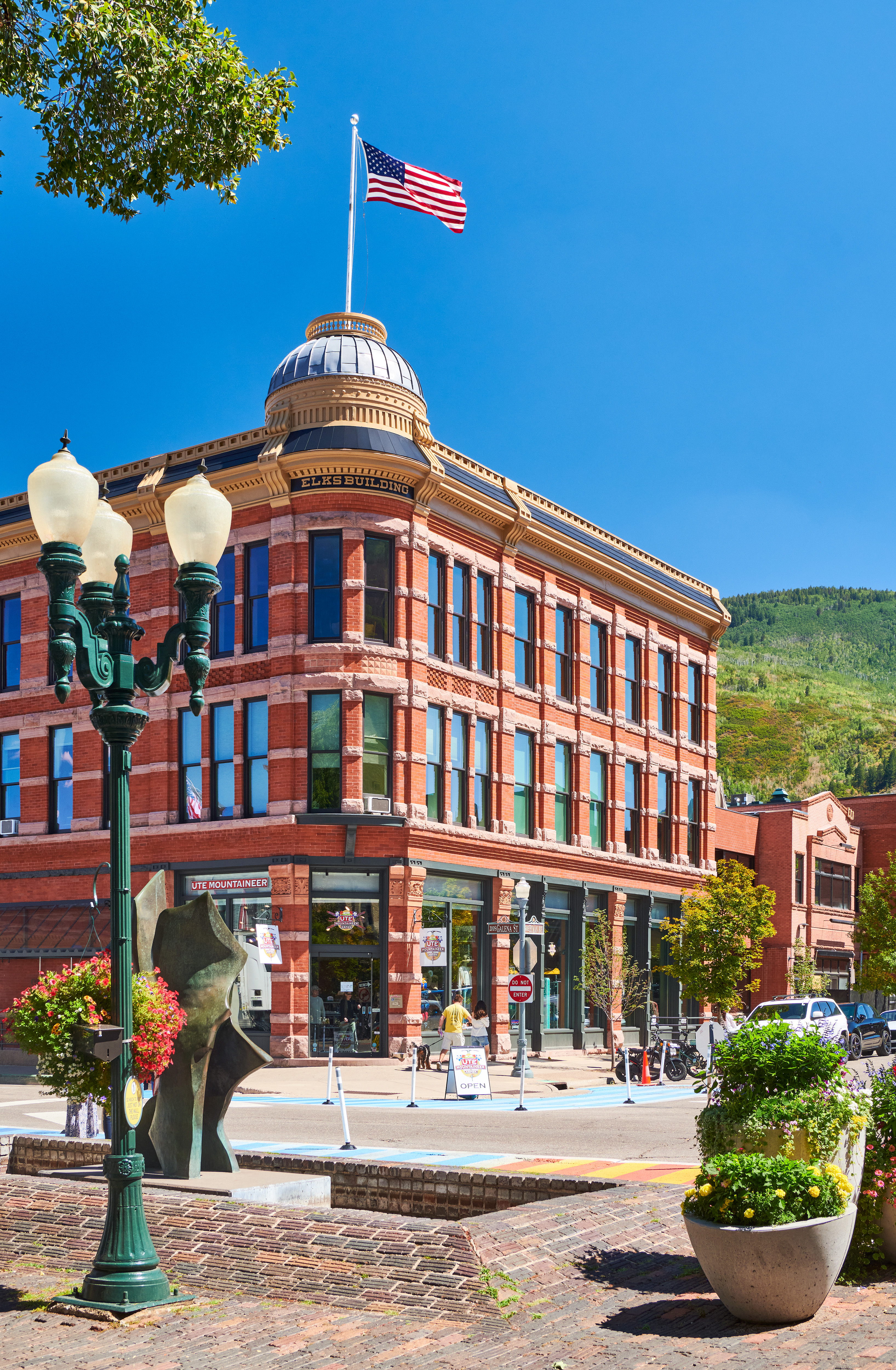 This iconic downtown building, known as the Elks Building, showcases turn-of-the-century brick architecture with a prominent domed corner tower. The ground level features retail space under the Ute Mountaineer sign, while the upper stories display classic arched window detailing. The property occupies a prime corner lot, enhanced by surrounding street furniture and vibrant landscaping, illustrating a high-visibility commercial asset in a historic setting.