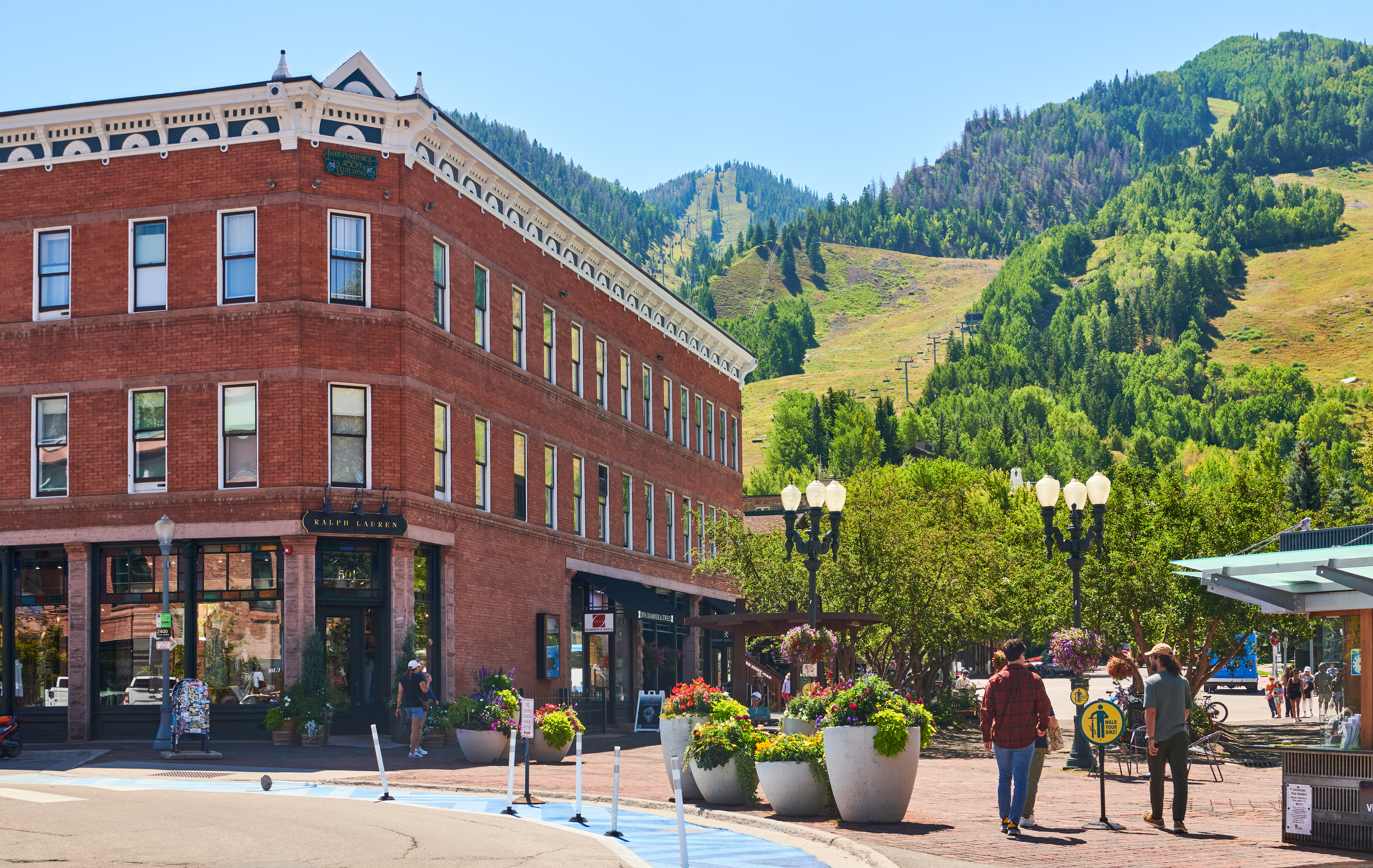 This image captures the historic Independence Building in a vibrant, pedestrian-friendly downtown setting. The prominent three-story brick structure features classic architectural details and houses commercial tenants, set against a stunning backdrop of a lush, mountainous ski resort landscape. The scene is enhanced by well-maintained planters, decorative street lamps, and a lively, accessible streetscape.