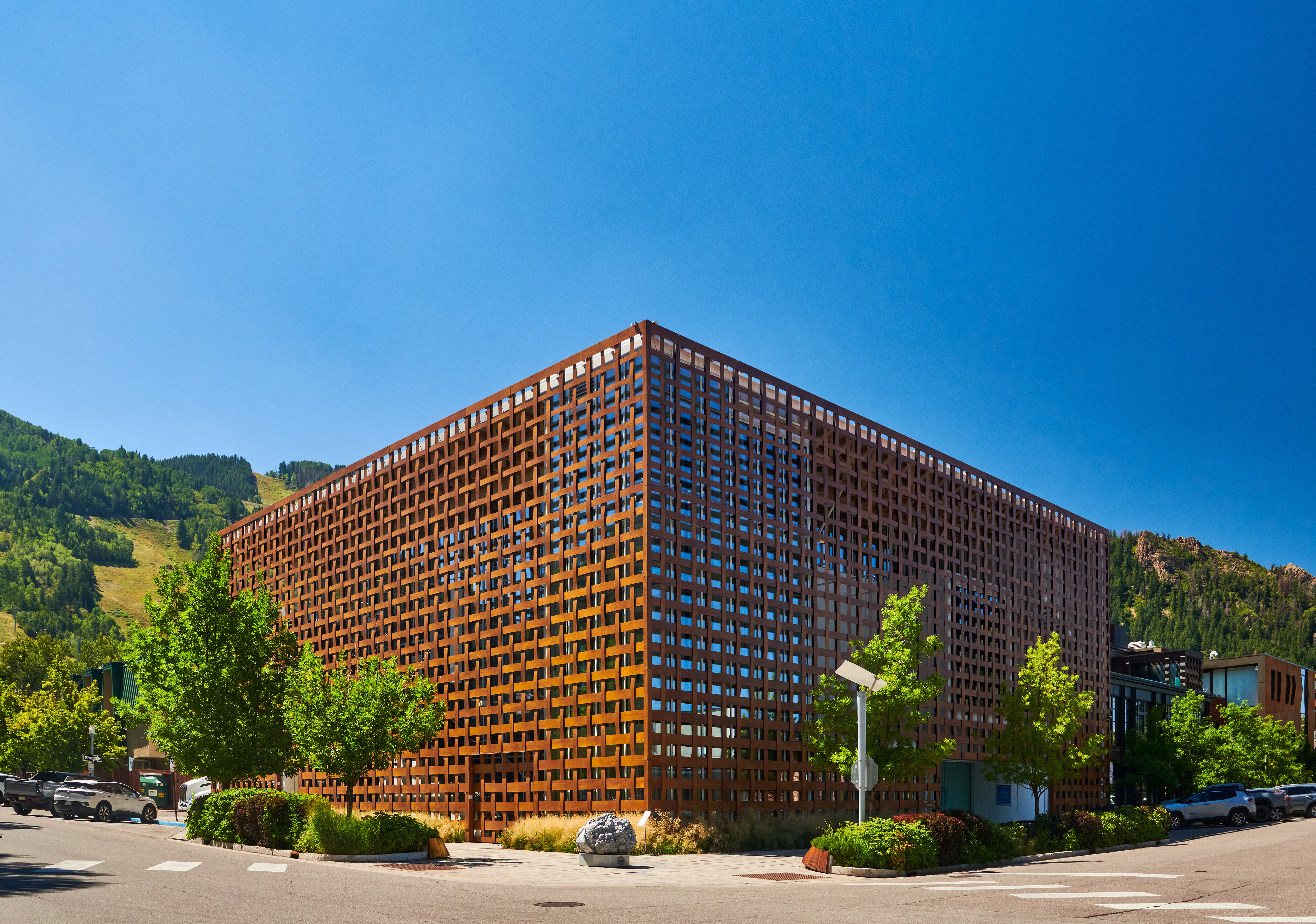 This unique commercial building features a striking, multi-layered Corten steel facade arranged in a woven, grid-like pattern that offers both privacy and architectural depth. The structure is set against a backdrop of mountains on a clear day, showcasing a modern, industrial aesthetic that stands out in the urban landscape. It is situated on a street corner, featuring professional landscaping and a clean, minimalist design approach.