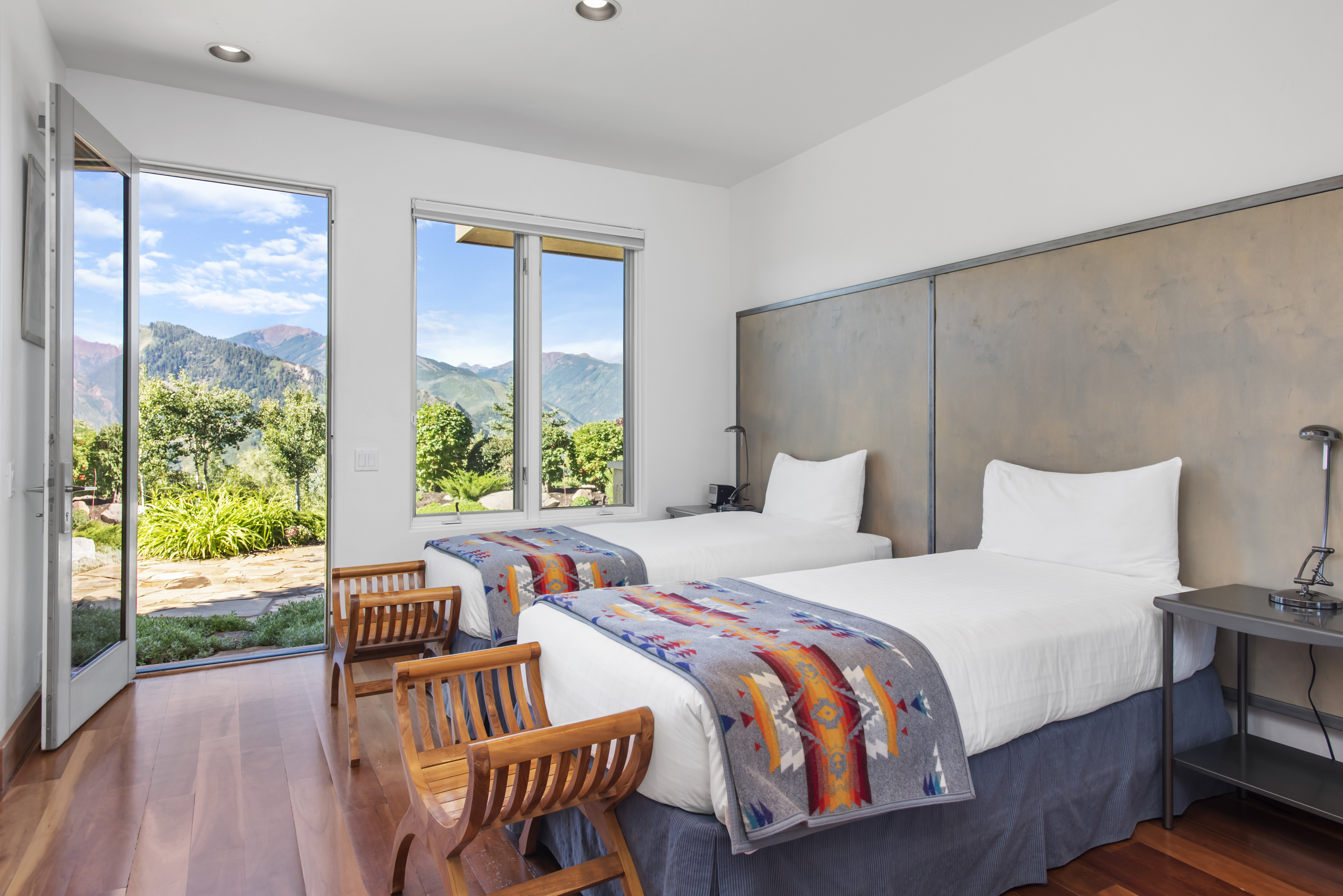 This guest bedroom features two twin-sized beds dressed with southwestern-style blankets, set against a sleek metallic headboard wall. Large windows and a glass door provide an abundance of natural light and direct access to a scenic patio with mountain views. The room is finished with warm hardwood flooring and rustic wood accent chairs, creating a cozy and inviting mountain retreat aesthetic.