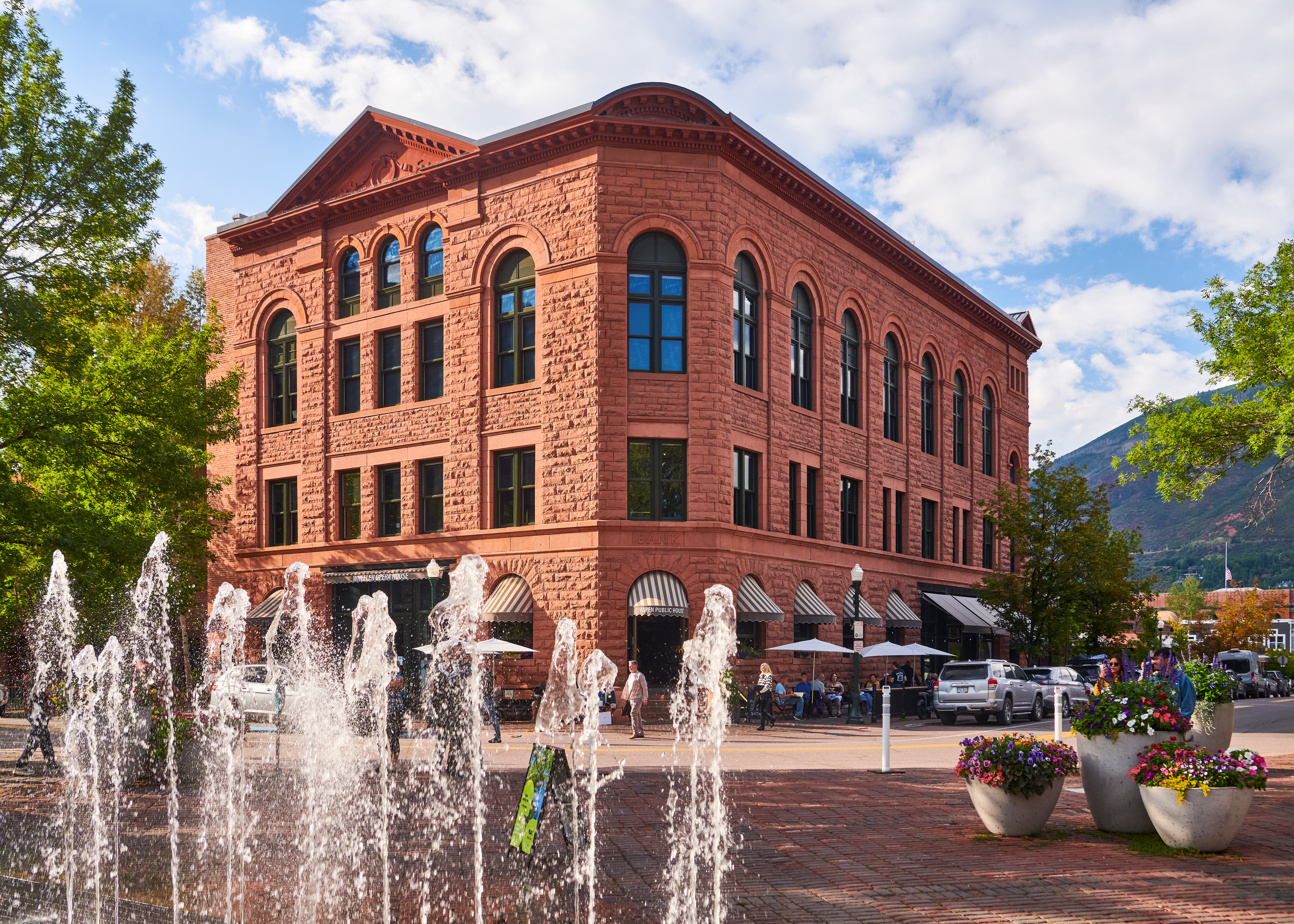 This stunning stone-clad building features classic Romanesque Revival architecture, characterized by its textured masonry, arched windows, and decorative cornice. Situated in a vibrant urban setting, it includes a prominent street-level entrance and outdoor patio seating, complemented by a public fountain in the foreground. Its historic charm and street-level commercial presence make it an iconic, high-visibility property.