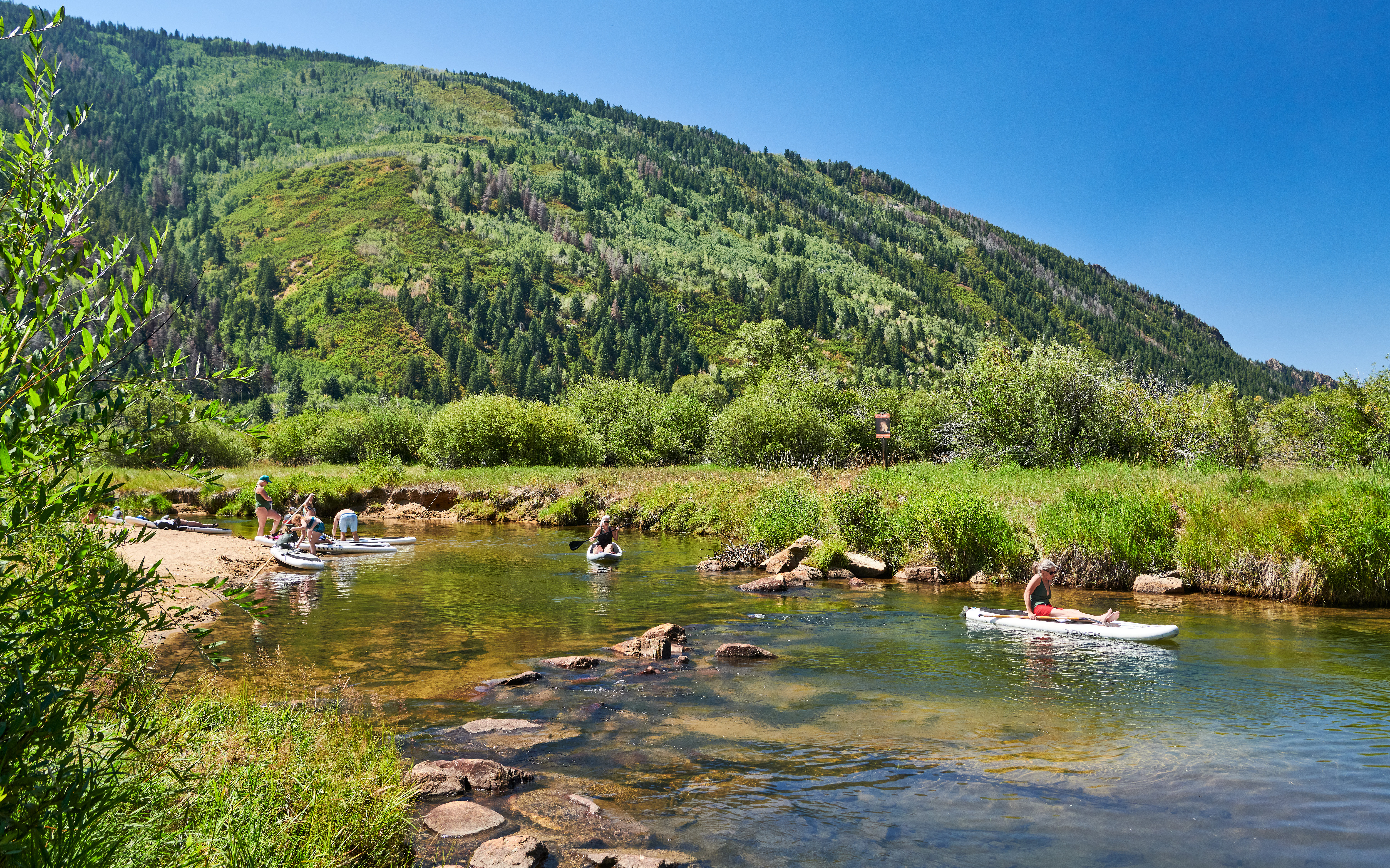 This scenic setting highlights a natural waterway suitable for recreational activities like paddleboarding, set against the backdrop of a sprawling mountain range covered in evergreen and deciduous forests. The landscape features a mix of lush grassy banks and rocky shallows, emphasizing the outdoor lifestyle and recreational appeal of the property's surrounding community environment.