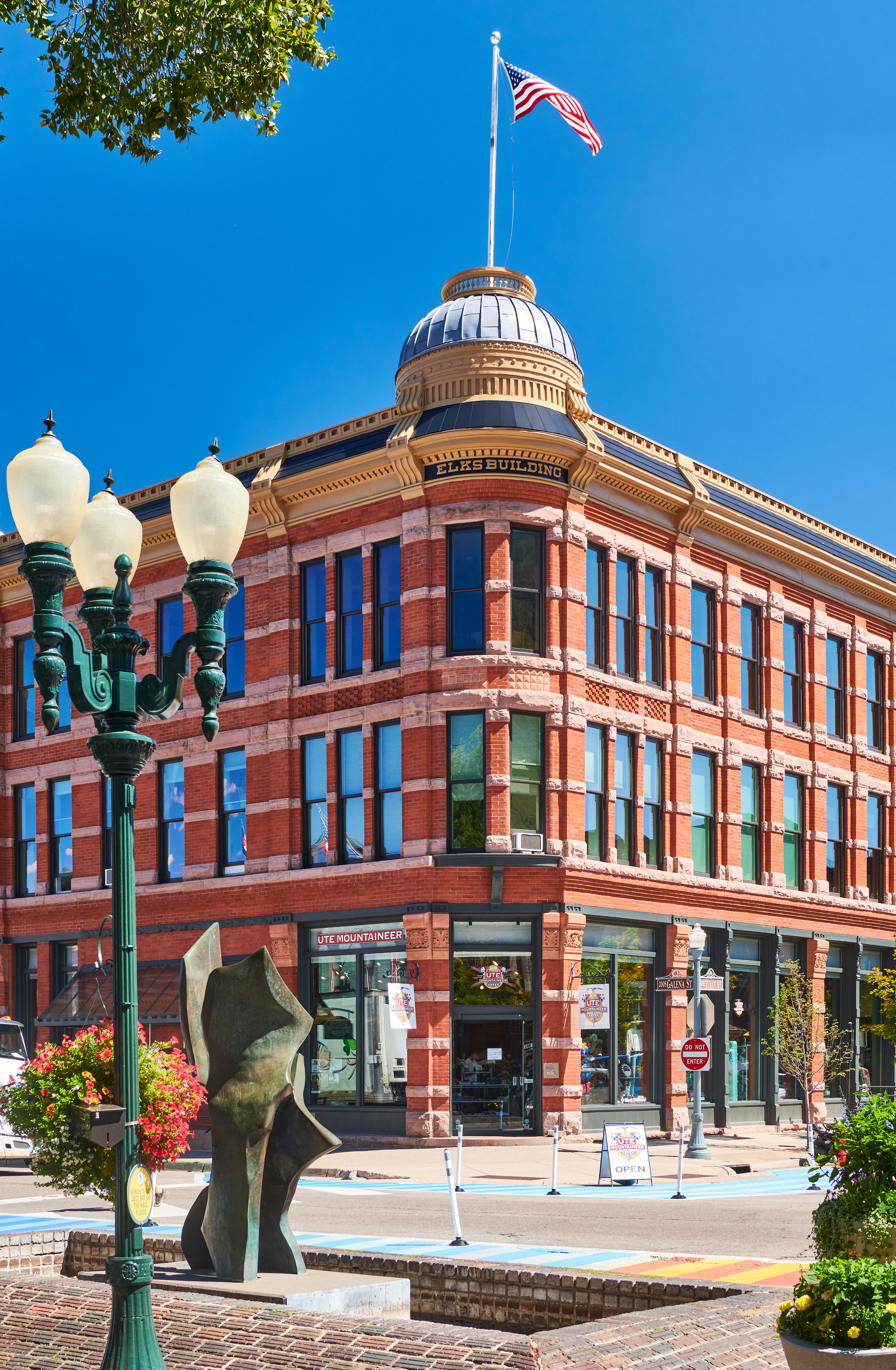 This stunning historical building, known as the Elks Building, prominently features classic red brick masonry with decorative stone accents and a distinctive corner dome. The street-level frontage offers prominent storefront retail space, complemented by decorative street lamps and a vibrant public plaza area. The architecture exudes a timeless, ornate charm characteristic of landmark downtown buildings.