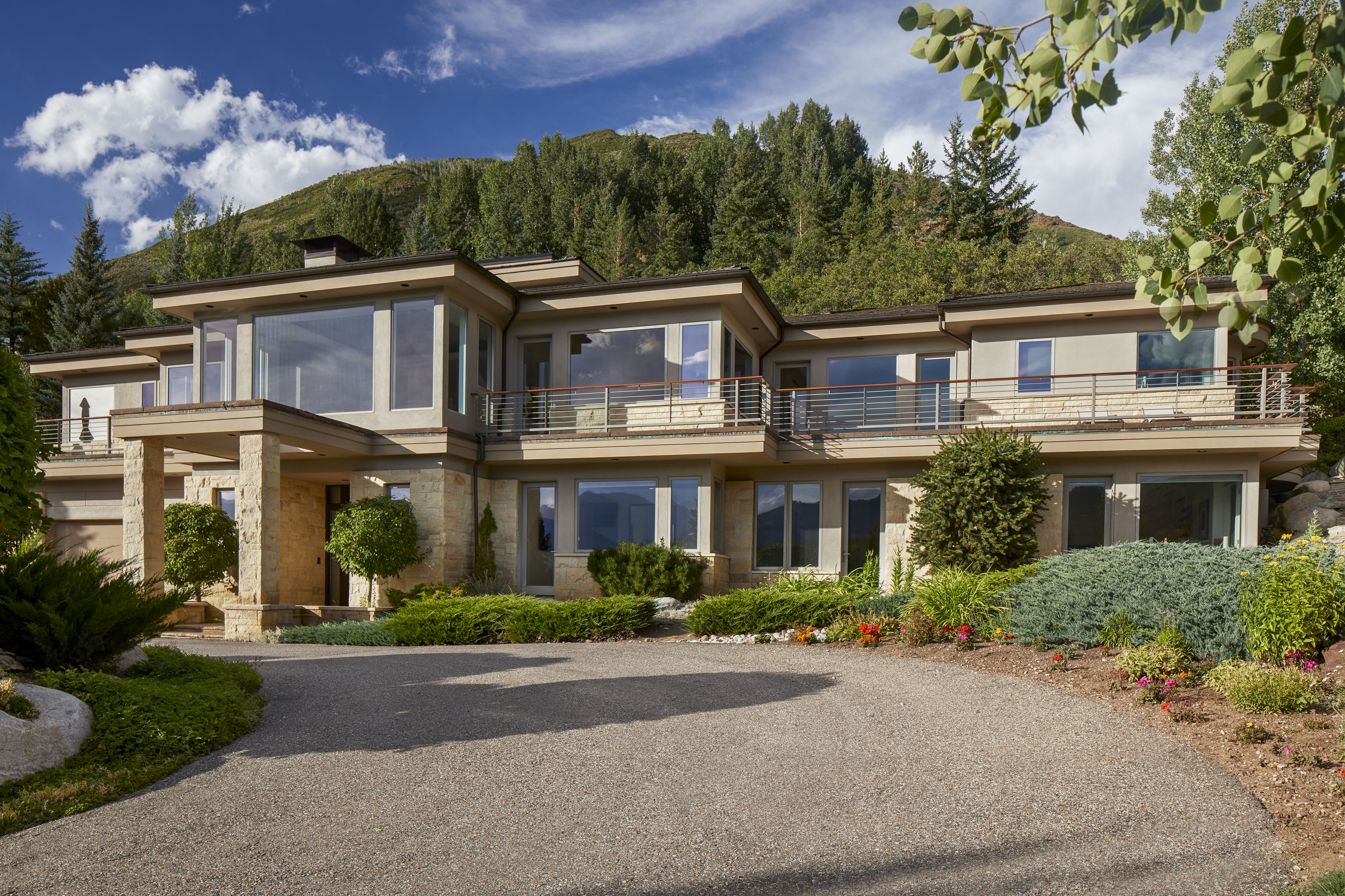 This striking multi-story luxury mountain residence features a blend of natural stone and smooth stucco siding, exuding a high-end, contemporary aesthetic. A spacious, elevated wrap-around balcony with sleek metal railings provides expansive views of the lush, tree-filled landscape and mountain backdrop. The property is complemented by a paved circular driveway and meticulously landscaped gardens, creating a sophisticated and welcoming approach to the home.