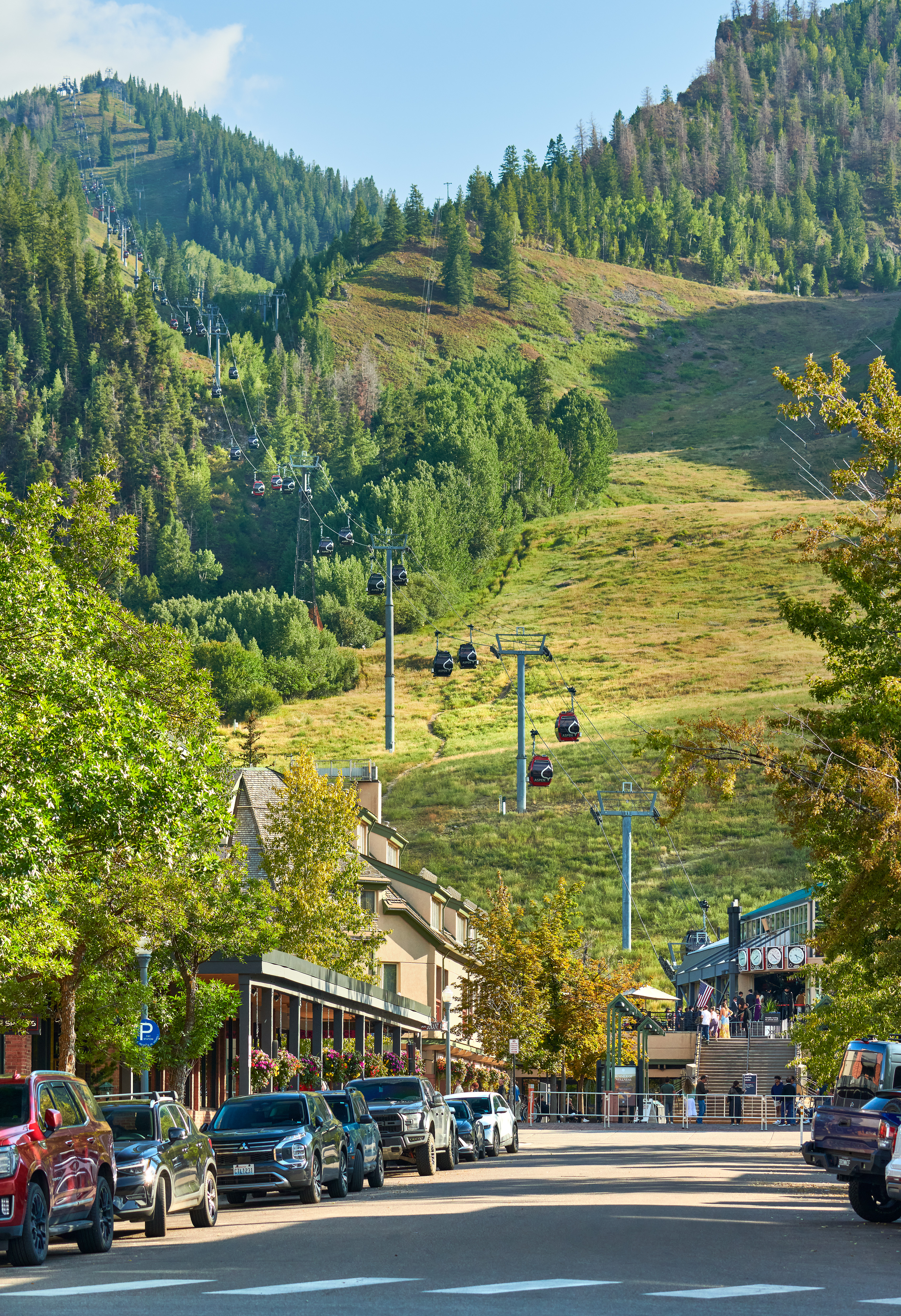 This image showcases a vibrant street scene at the base of the Aspen Mountain ski resort, highlighting its prime location near the Silver Queen Gondola. The setting captures the bustling atmosphere with close proximity to dining, retail, and recreational access, framed by scenic mountain slopes. This area represents highly desirable mountain-town living, emphasizing walkability and direct access to year-round outdoor amenities.
