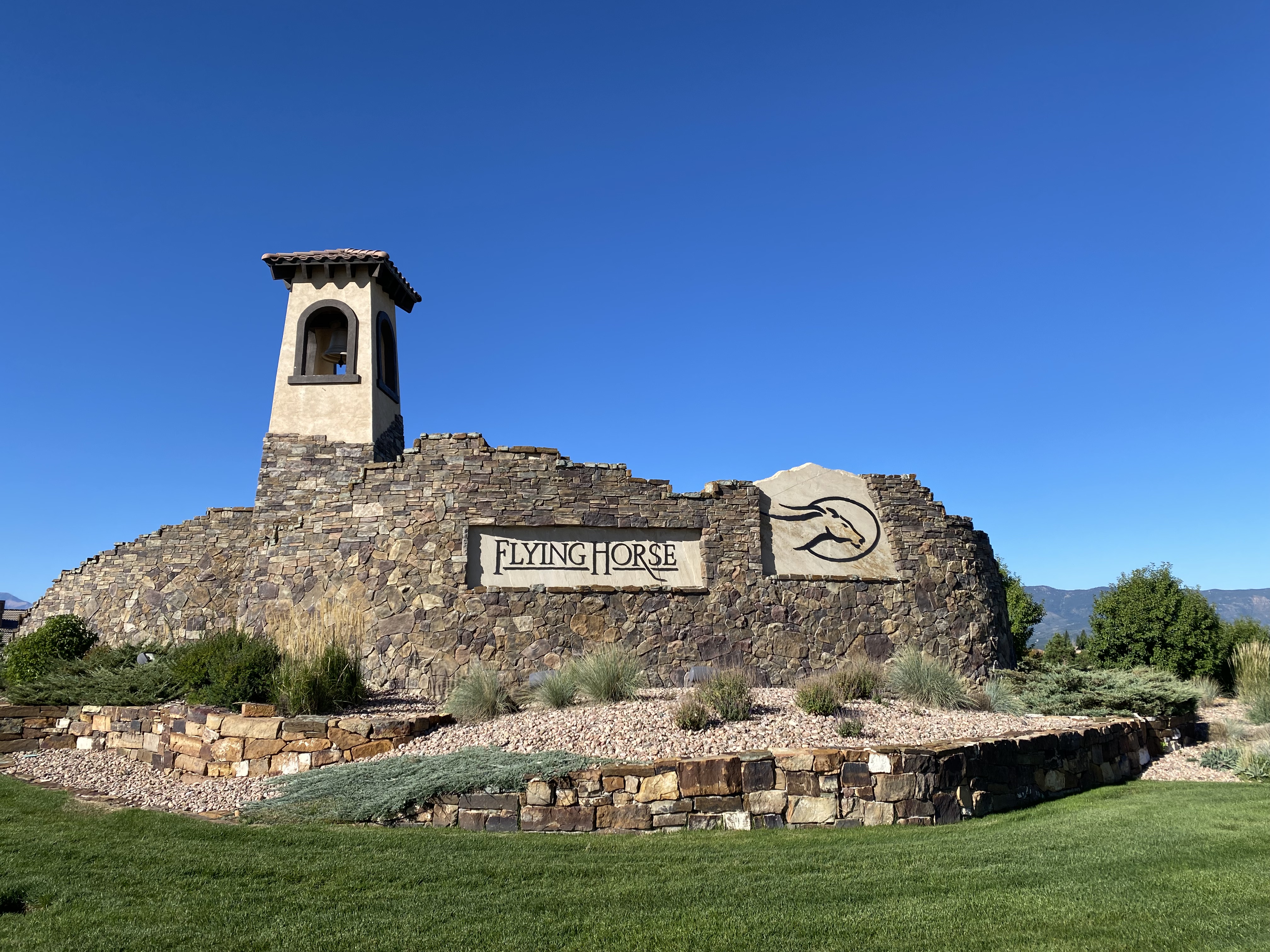 This image captures the prominent stone monument sign for the Flying Horse community, featuring a distinctive bell tower structure and a stylized horse logo. The sign is set against a backdrop of a clear blue sky and manicured landscaping, with a low stone retaining wall framing the foreground. The perspective is a wide, eye-level shot that emphasizes the grand, rustic aesthetic of the neighborhood entrance.