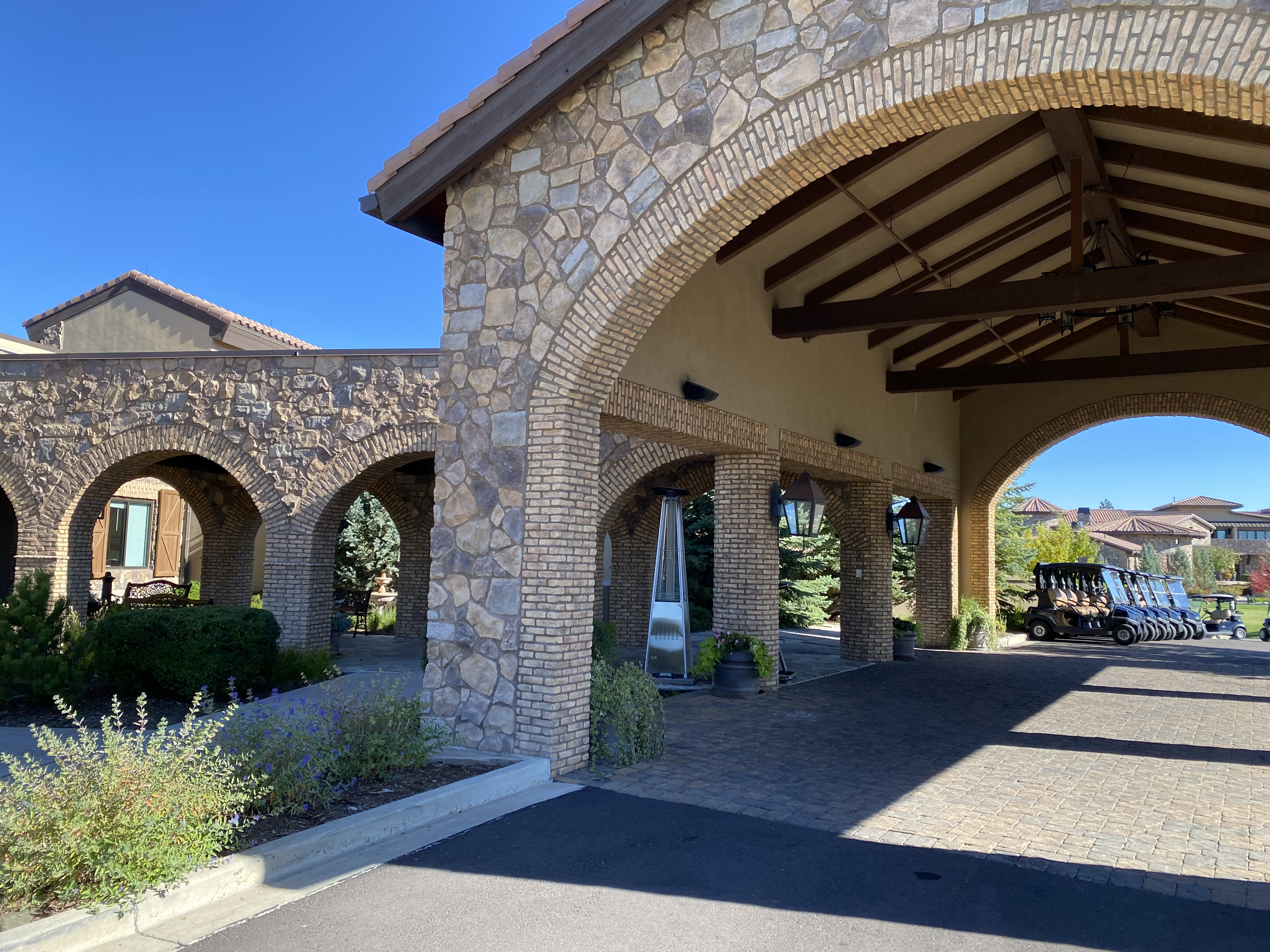 This image captures the grand porte-cochère entrance of a clubhouse, characterized by its rustic Mediterranean architecture featuring stone-clad columns and arched brick detailing. The covered driveway provides a sheltered arrival point, leading the eye toward a row of golf carts parked in the distance under a clear blue sky. The scene conveys a sense of upscale leisure and professional maintenance, typical of a high-end golf course or resort facility.