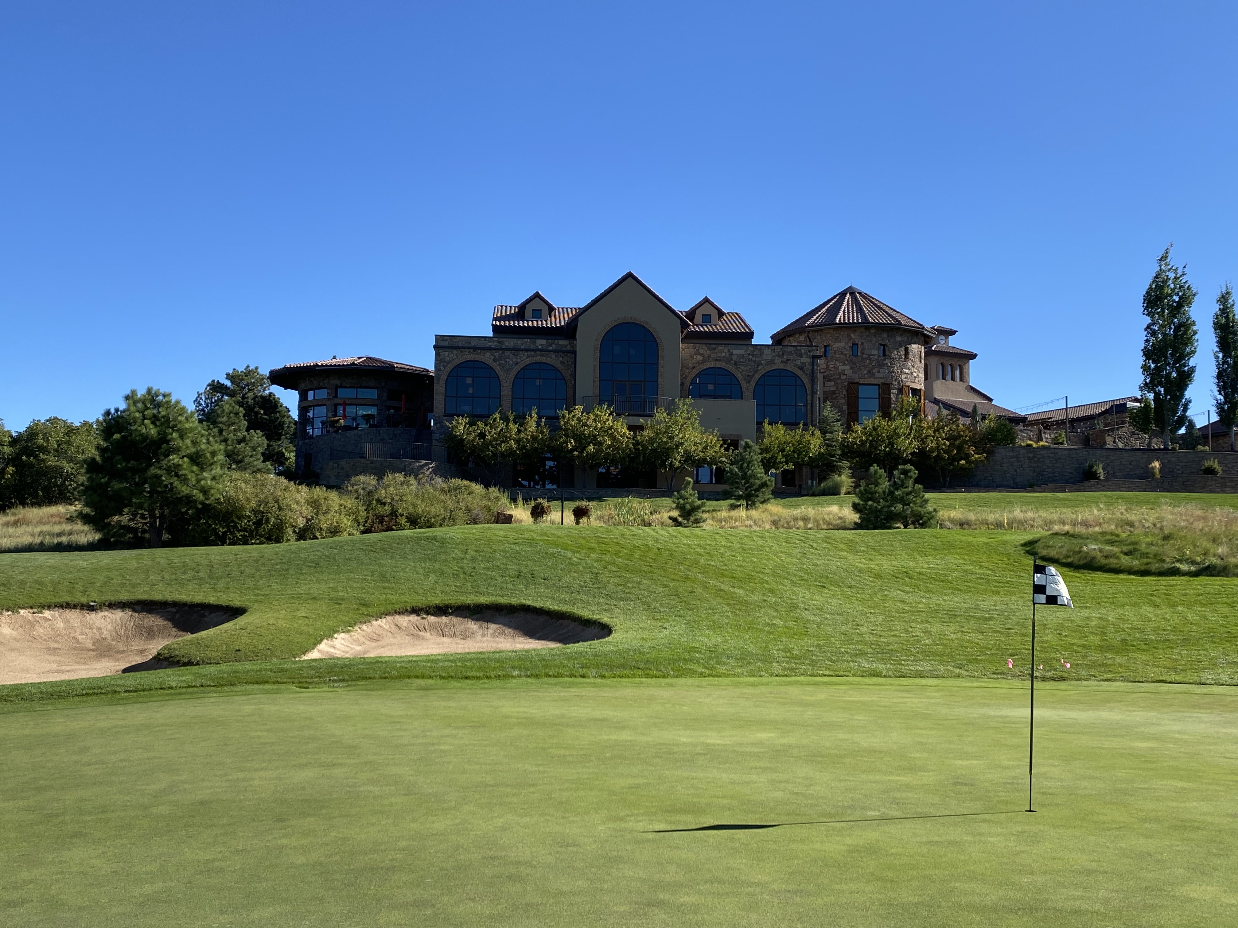 This image captures a sprawling, Mediterranean-inspired clubhouse overlooking a meticulously manicured golf course. The structure features a complex roofline with multiple gables and a prominent circular tower, all set against a clear blue sky. In the foreground, a putting green with a flagstick leads the eye toward the building, emphasizing the property's recreational appeal and expansive outdoor space.
