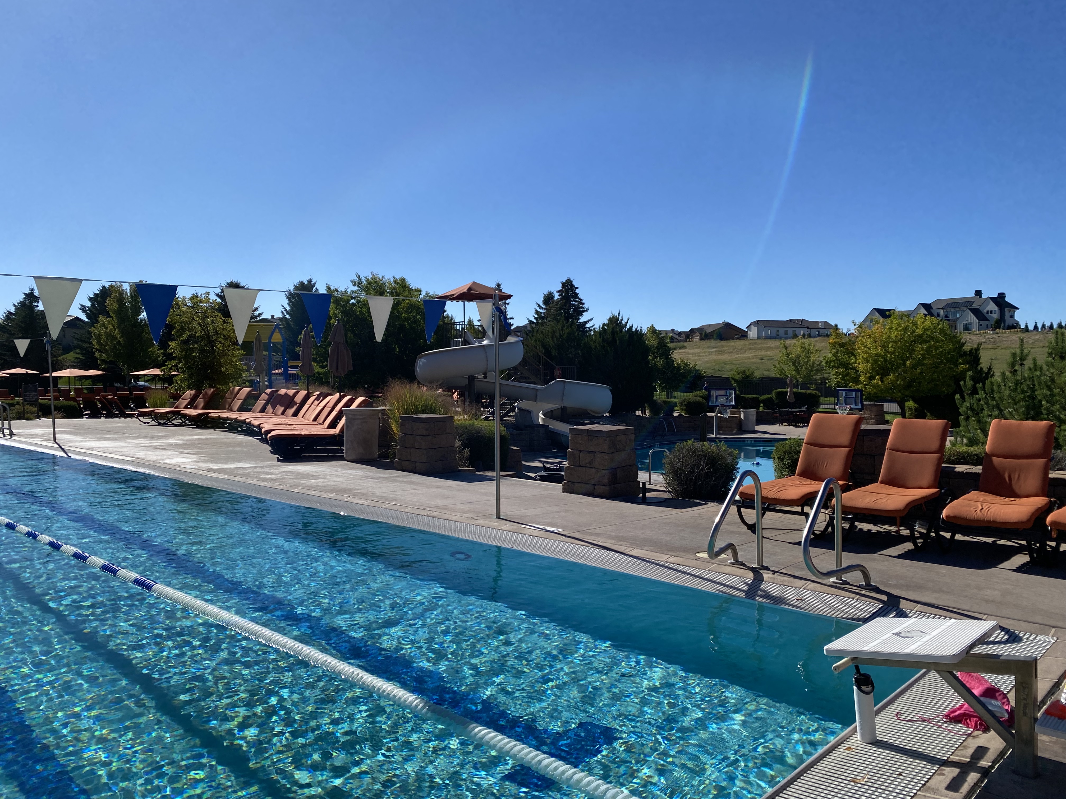 This outdoor community pool area features a long lap pool with a lane divider and a starting block in the foreground. The pool deck is lined with numerous orange lounge chairs, a water slide, and decorative pennant flags strung across the space. The background showcases a scenic, grassy hillside with residential homes, creating a bright and inviting recreational atmosphere.