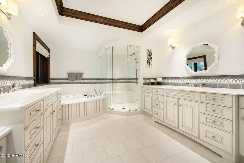 This is a spacious primary bathroom featuring dual vanities with white cabinetry and marble countertops. A glass-enclosed shower and a separate soaking tub are also present. The room is well-lit and has a clean, luxurious feel, enhanced by the neutral color palette and tile flooring.