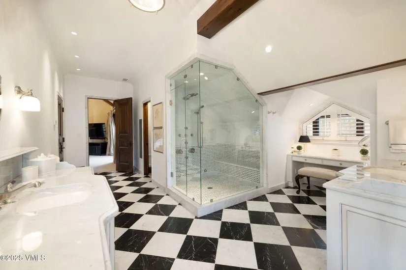 This is a primary bathroom featuring a black and white checkered floor, a glass-enclosed shower with white tile, and a vanity area with a window. The bathroom has a luxurious and classic feel, with white walls and marble countertops. The perspective is from the doorway, showcasing the entire space.