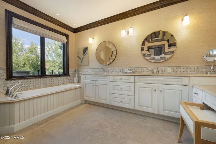 This is a well-lit primary bathroom featuring a double vanity with white cabinetry and light countertops. Two round mirrors hang above the sinks, complemented by sconce lighting. A large window provides natural light above a soaking tub, and the overall design is elegant and traditional.