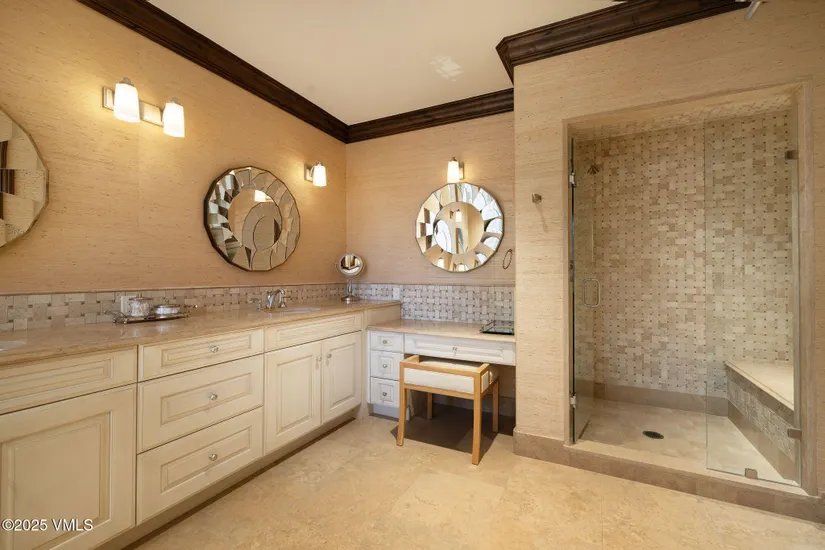 This is a well-lit primary bathroom featuring a long vanity with light-colored cabinetry and a beige countertop. Two decorative round mirrors hang above the sinks, complemented by sconce lighting. A glass-enclosed shower with tiled walls is visible on the right, and a small vanity table with a stool is positioned next to the main vanity, creating a luxurious and functional space.