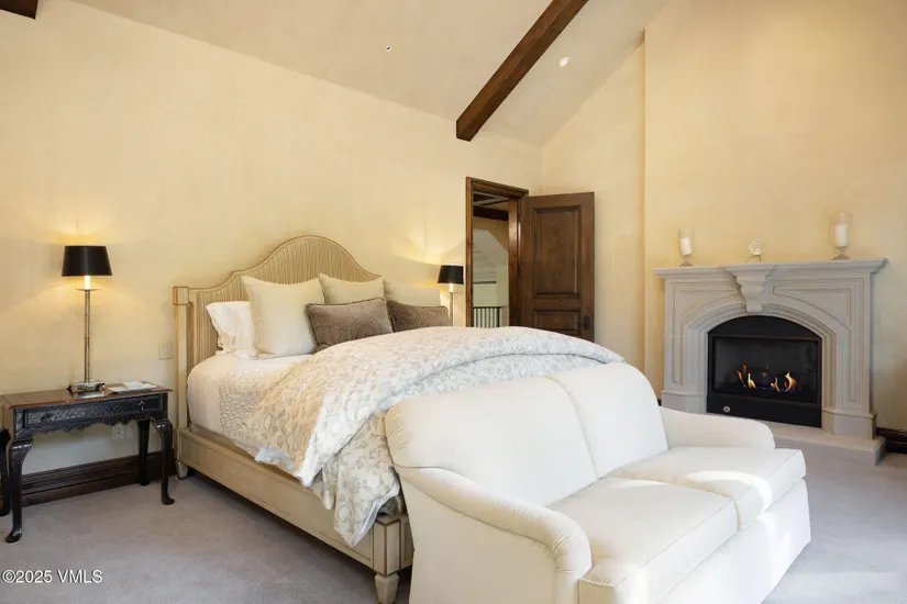 This is a primary bedroom featuring a bed with a decorative headboard, neutral-toned bedding, and a small sofa at the foot of the bed. A fireplace with a decorative mantel adds a cozy focal point. The room is painted in warm, inviting colors, and a dark wood nightstand with a lamp sits beside the bed.