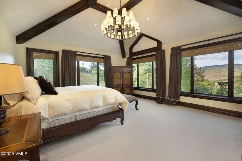 This is a primary bedroom featuring a king-size bed with white linens and a dark brown accent pillow. The room has a vaulted ceiling with exposed dark wood beams and a large chandelier. There are multiple windows with dark wood frames and brown curtains, offering a view of the surrounding landscape. A dark wood armoire is also visible.
