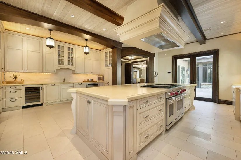 This is a well-lit kitchen featuring custom white cabinetry, a large central island with a light-colored countertop, and high-end appliances. The ceiling has exposed wooden beams, and the flooring is light tile. The kitchen has a luxurious and spacious feel, perfect for entertaining.