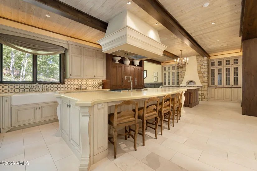 This is a spacious kitchen featuring a large island with seating, custom cabinetry, and a light-colored countertop. The kitchen has a rustic yet elegant feel with wooden beams on the ceiling and a stone accent wall with a pizza oven. The flooring is light tile, and natural light streams in through a window above the sink.