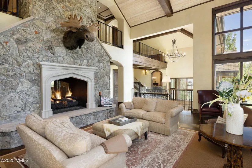 This is an interior shot of a luxurious living room featuring a stone fireplace with a mounted moose head. The room is spacious with high ceilings, exposed beams, and a second-floor balcony overlooking the area. The decor includes comfortable sofas, a patterned rug, and elegant lighting, creating a warm and inviting atmosphere.