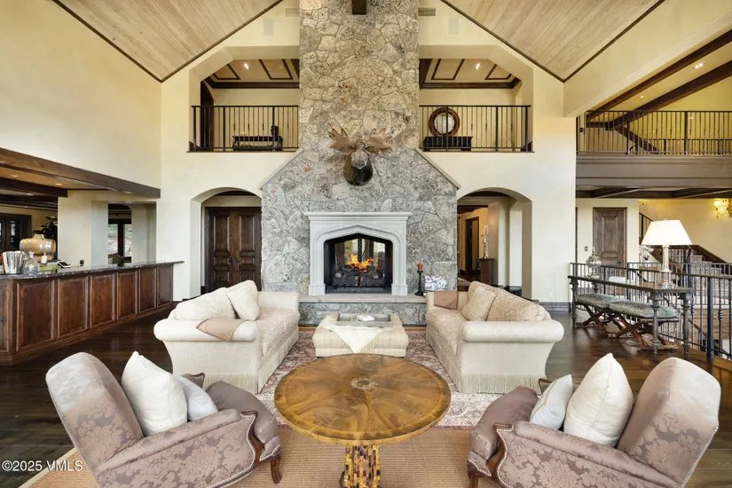 This grand living room features a soaring ceiling, a massive stone fireplace with a mounted moose head, and two balconies overlooking the space. Two sofas and two armchairs are arranged around a round coffee table on a patterned rug, creating a luxurious and inviting atmosphere. The room is bathed in natural light, highlighting the rich wood floors and architectural details.