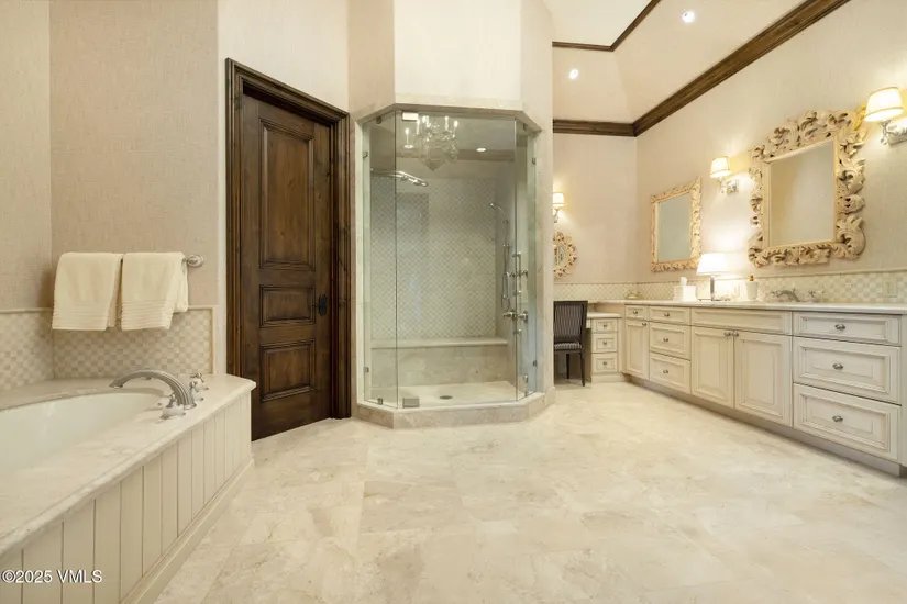 This is a luxurious primary bathroom featuring a large, glass-enclosed shower, a soaking tub, and a double vanity with ornate mirrors. The room is finished with light-colored marble flooring and neutral wall tones, creating a serene and elegant atmosphere. A dark wood door adds a touch of contrast and sophistication.