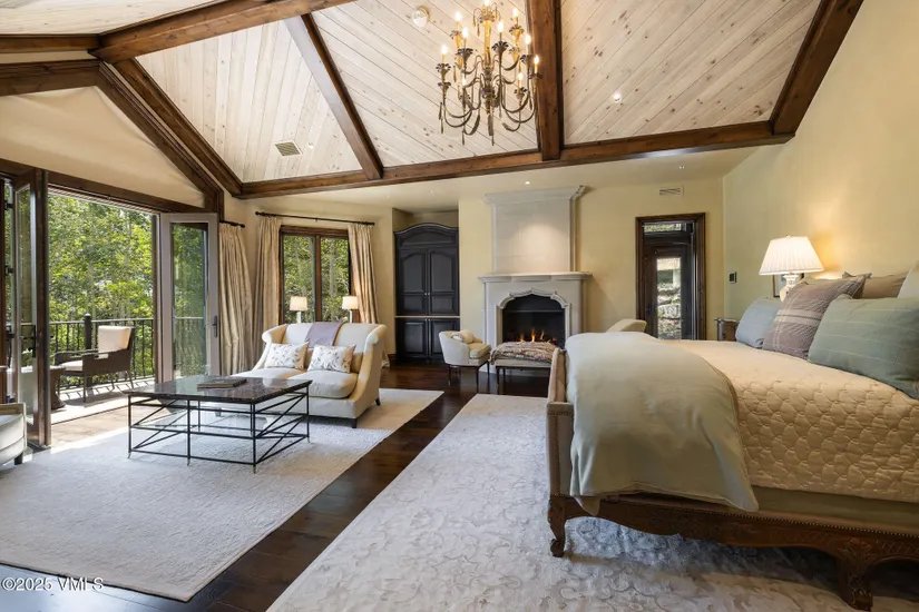 This is a luxurious primary bedroom featuring a high vaulted ceiling with exposed wooden beams and a grand chandelier. The room includes a fireplace, a seating area with a sofa, and access to a balcony through large glass doors, offering a seamless indoor-outdoor living experience. The color palette is warm and inviting, creating a serene and elegant atmosphere.