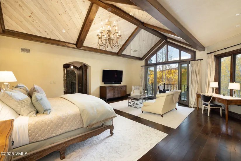 This is a luxurious primary bedroom featuring a vaulted ceiling with exposed wooden beams and a decorative chandelier. The room includes a large bed, a seating area with a chaise lounge, a desk, and access to a balcony through sliding glass doors. The color palette is warm and inviting, creating a comfortable and elegant atmosphere.