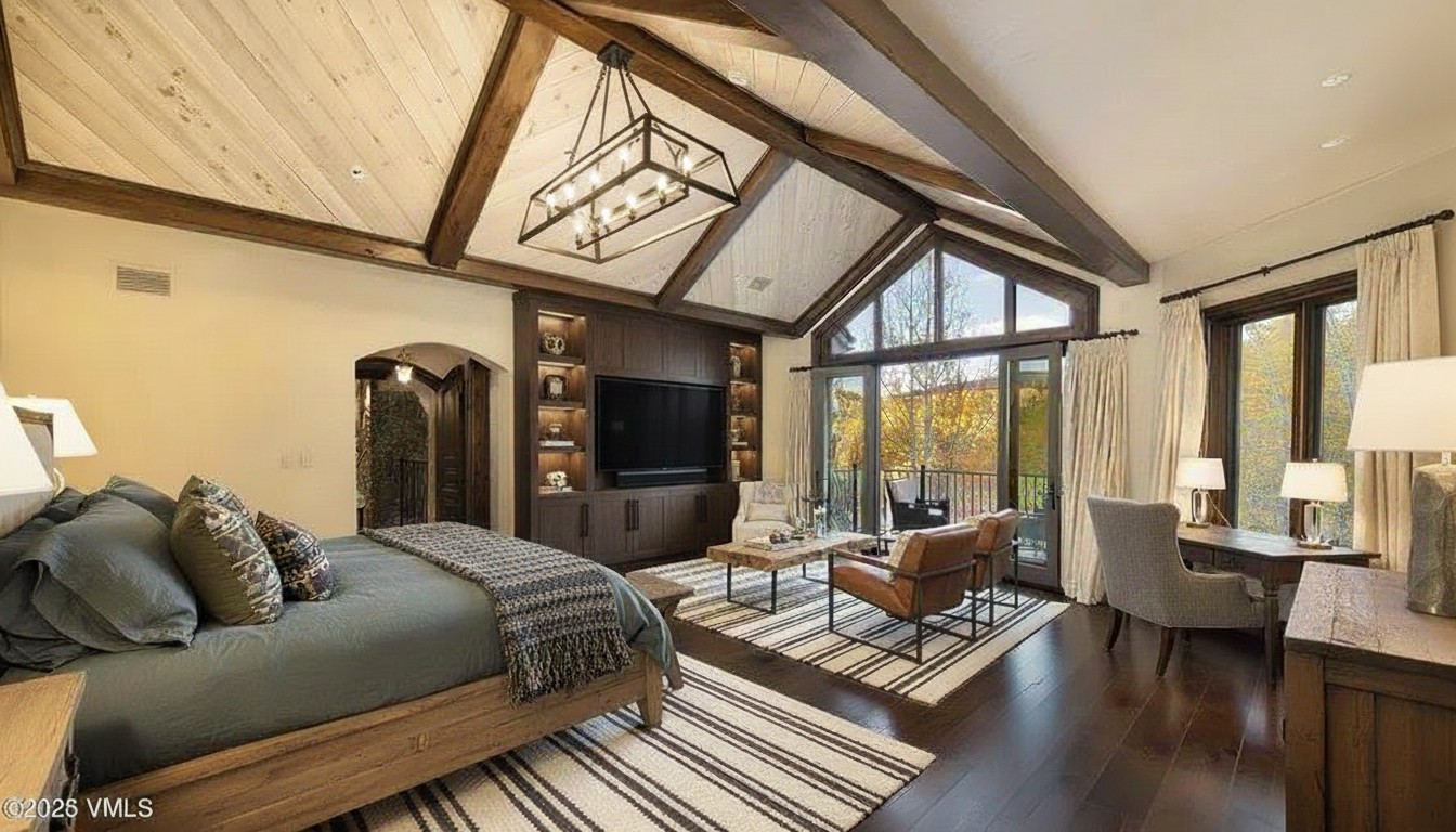 This is a primary bedroom featuring a vaulted ceiling with exposed beams and a modern chandelier. The room includes a built-in entertainment center, a king-size bed with a cozy throw, and a seating area with leather chairs. A balcony is accessible through glass doors, offering natural light and views of the outdoors.