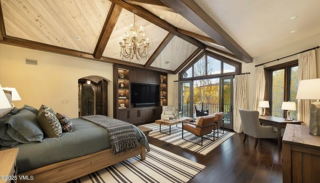 This is a primary bedroom featuring a vaulted ceiling with exposed wooden beams and a large chandelier. The room includes a bed with a wooden frame, a built-in entertainment center with a television, and a seating area with leather chairs. A balcony is accessible through glass doors, and a desk area is situated near a window, creating a luxurious and comfortable space.