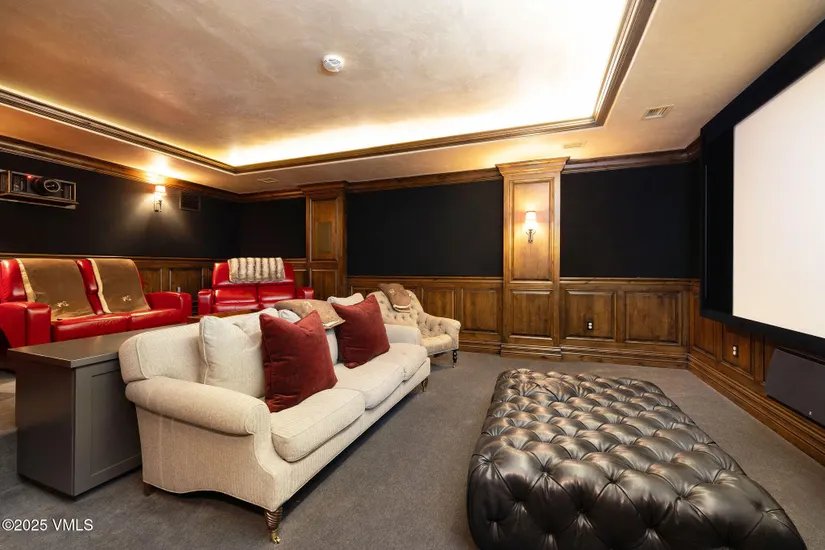 This is a luxurious home theater room featuring comfortable seating, including red leather recliners and a beige sofa with red pillows. The room has dark wood paneling and a large screen, creating a cozy and immersive viewing experience. A large leather ottoman sits in the foreground.