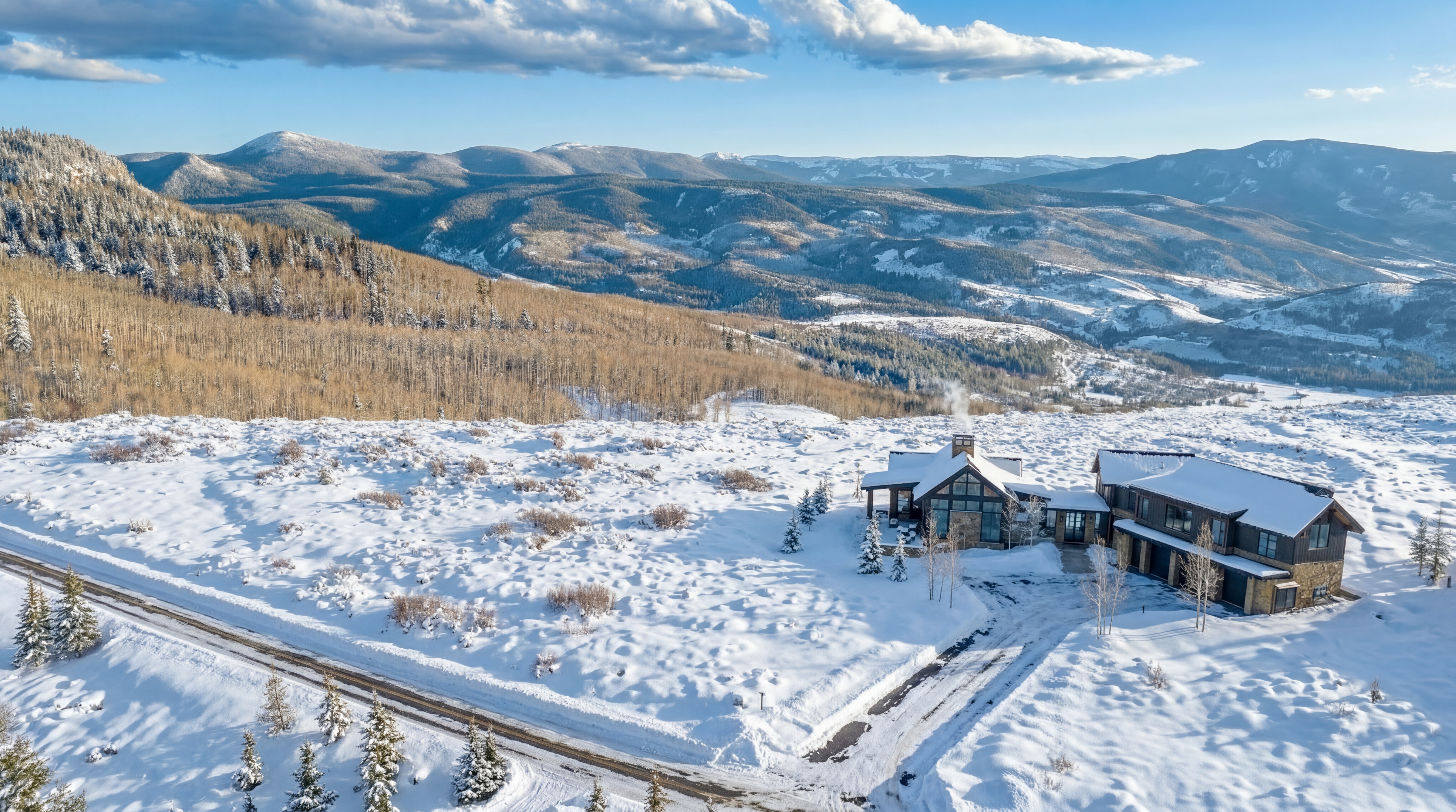 This is an impressive aerial view of a luxurious, modern mountain estate nestled in a snow-covered alpine landscape. The residence features high-end architectural elements like gabled roofs, stone masonry, and extensive windows that frame the expansive, panoramic mountain vistas. The property is well-integrated into its rugged, serene setting, highlighting a premium primary or secondary home in an exclusive, snowy environment.