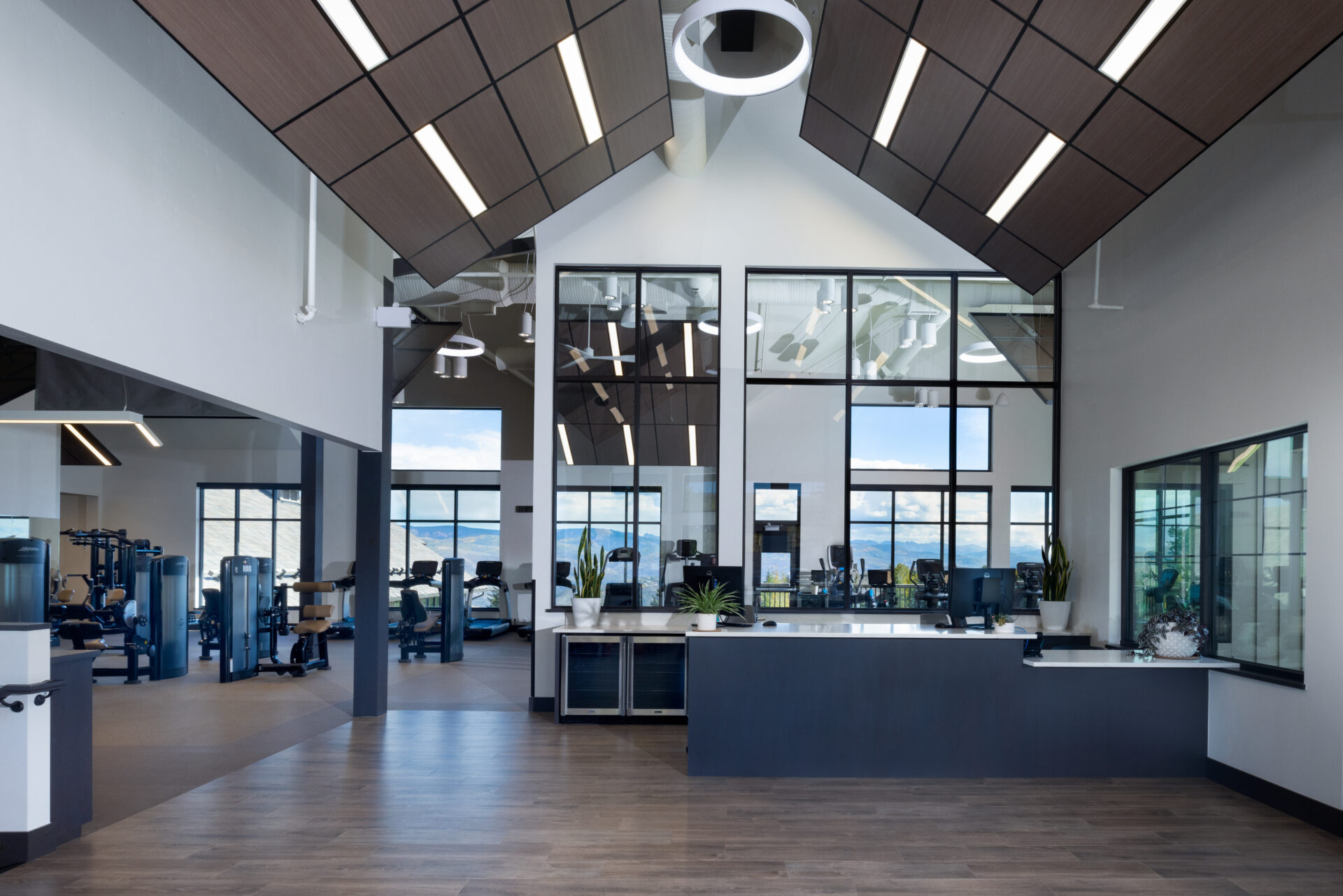 This gym or clubhouse reception area features a vaulted, wood-paneled ceiling with sleek linear lighting and a modern, minimalist reception desk. Large, floor-to-ceiling windows provide an abundance of natural light and scenic mountain views, while the adjacent fitness equipment indicates a well-equipped, high-end facility. The overall atmosphere is professional, airy, and contemporary, reflecting a resort-style amenity space.