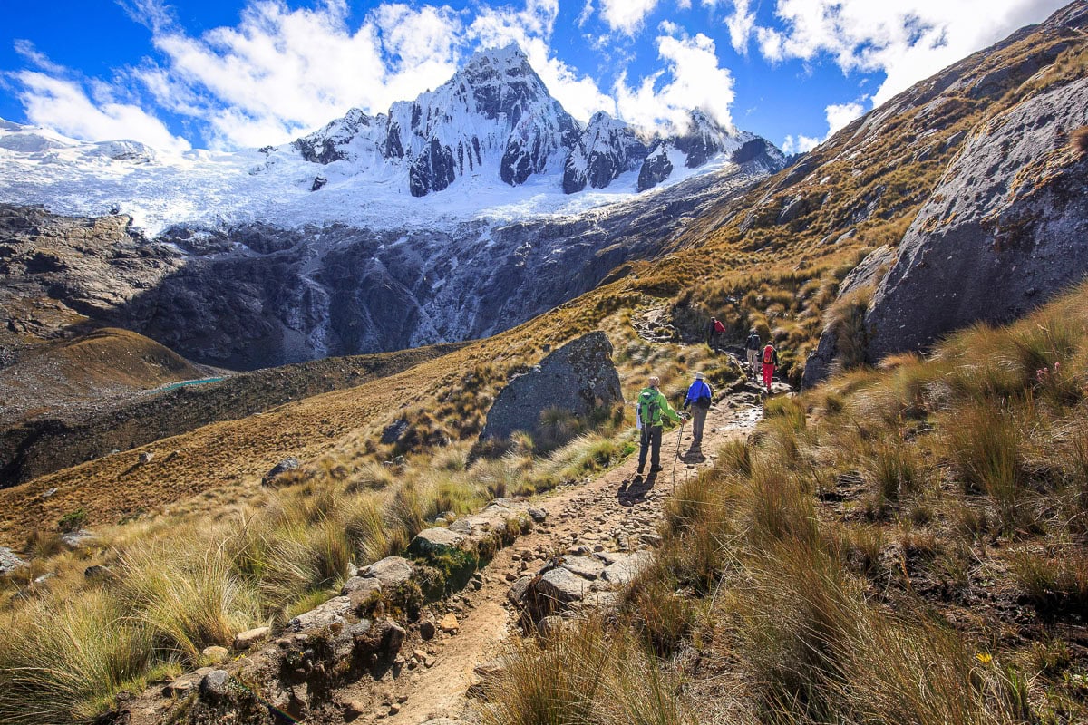 This image showcases a breathtaking mountain landscape featuring a rugged hiking trail winding through alpine terrain towards a prominent snow-capped peak. The scene is characterized by distinct grassy slopes, rocky formations, and a dramatic cloud-filled sky, conveying a sense of vast wilderness and outdoor recreational appeal. It serves as an example of scenic natural surroundings rather than a traditional residential property.