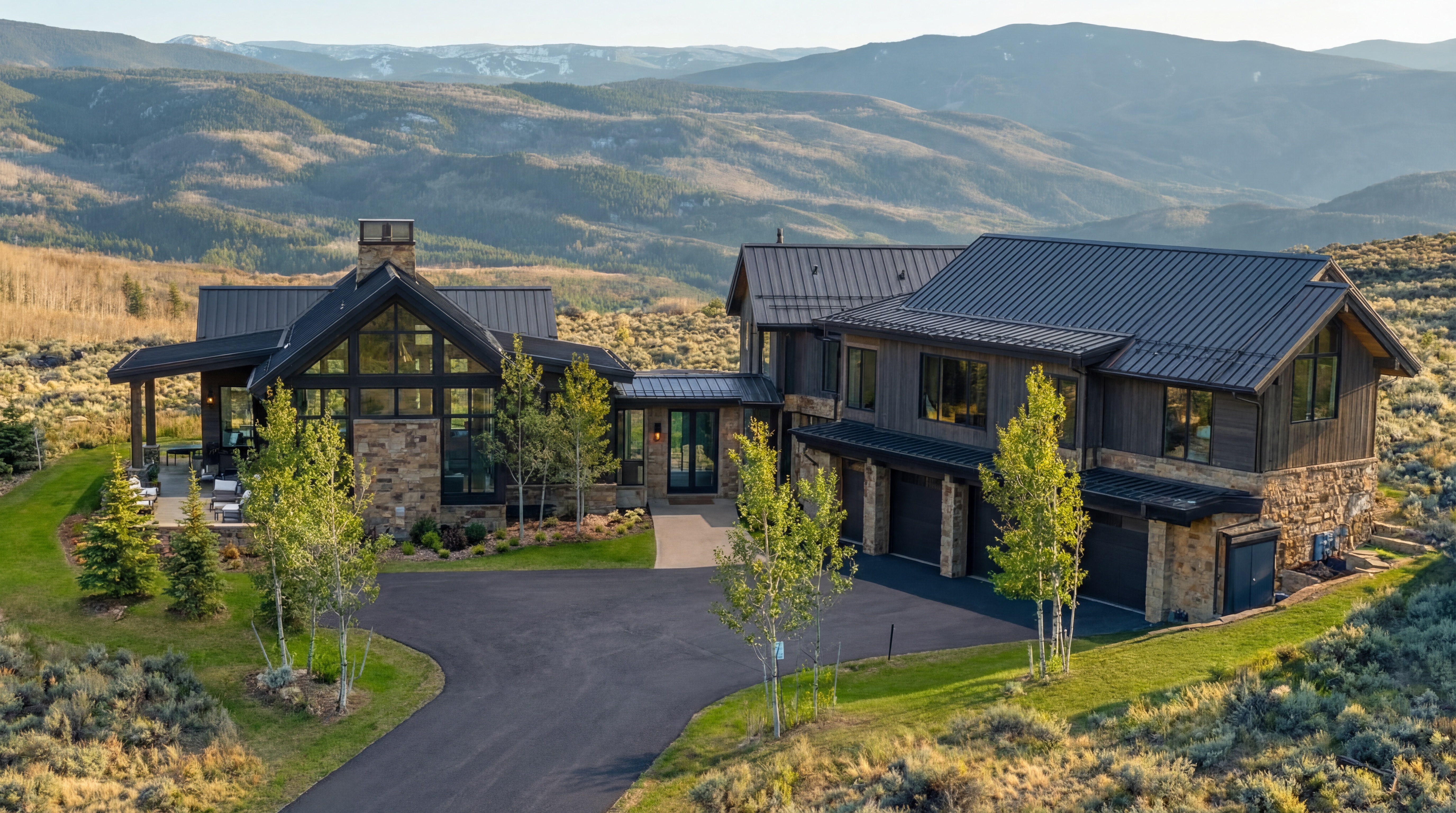 This stunning contemporary mountain estate features a harmonious blend of natural stone and dark timber siding, topped with sleek metal roofing. The expansive aerial perspective showcases the home's impressive architectural design, large framed windows, integrated garage, and well-manicured landscaping perfectly nestled against a vast, scenic mountain range backdrop. This property offers a luxurious, rustic-modern feel, ideal for those seeking a serene retreat.