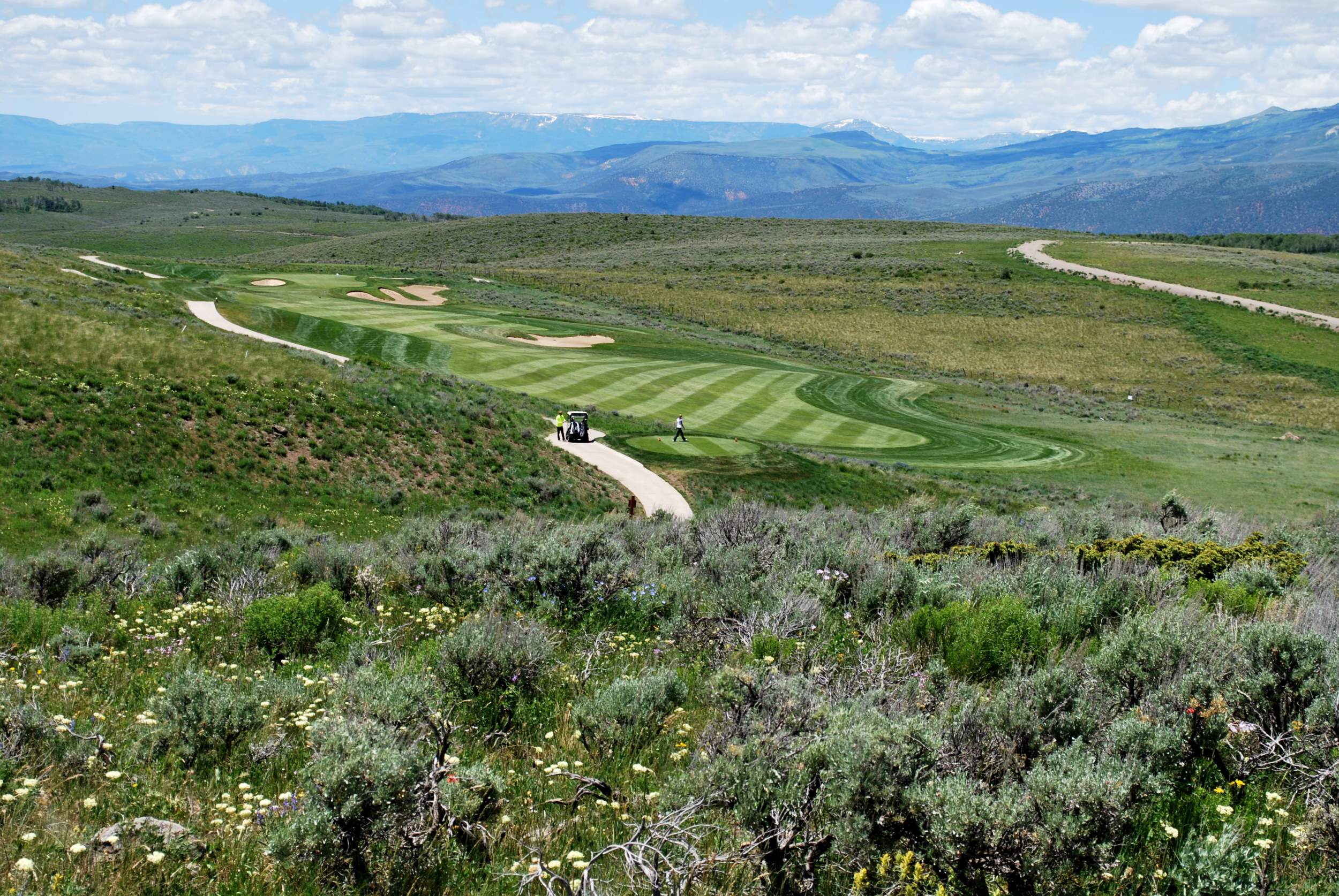 This image showcases a scenic golf course set against a backdrop of rolling mountains and expansive high-desert terrain. The course features meticulously maintained, patterned fairways that wind through natural landscape, emphasizing luxury resort-style recreation. It presents an appealing, serene environment perfect for those who value outdoor amenities and natural beauty in a property location.