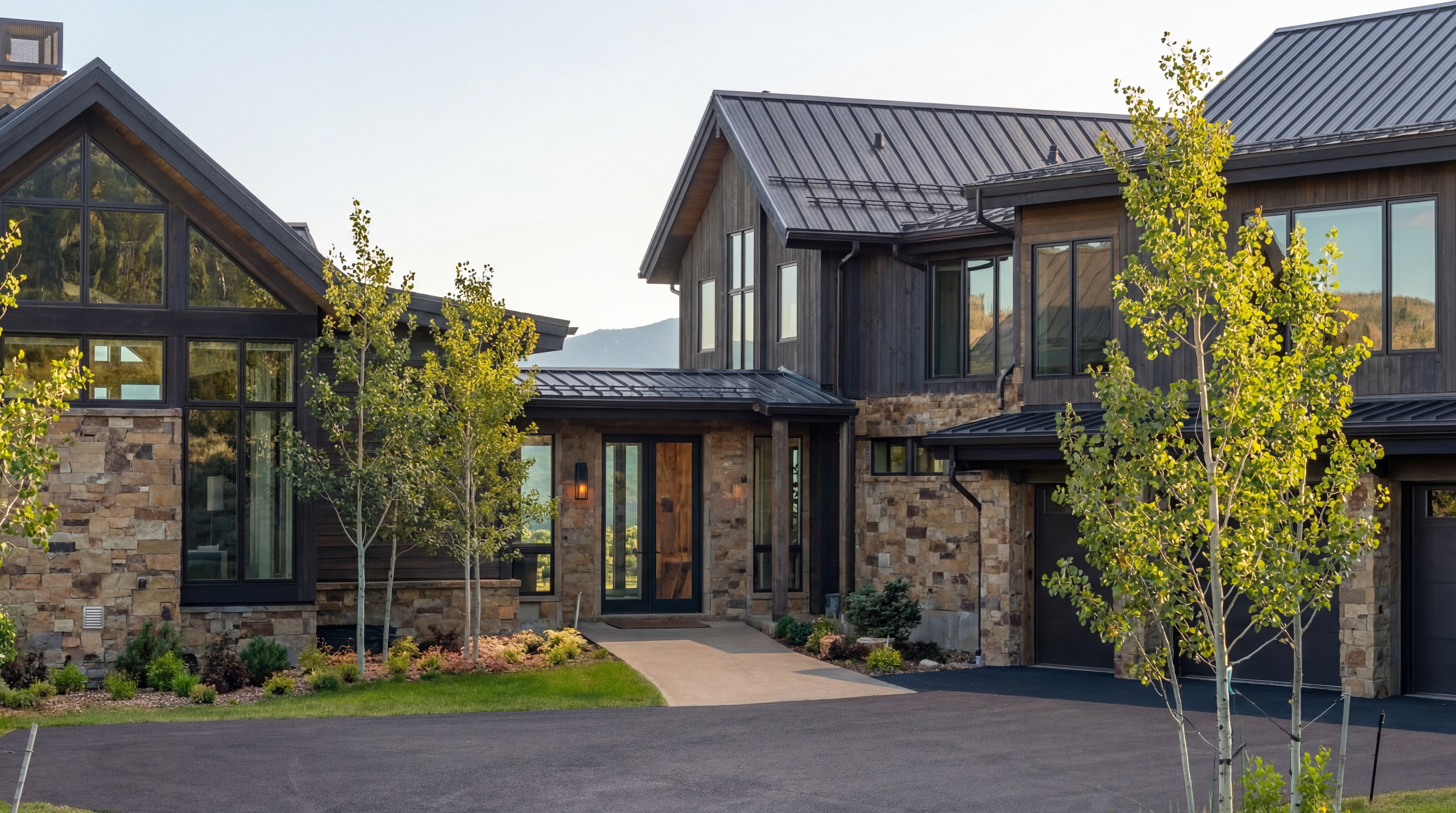 This stunning contemporary mountain home features a sophisticated blend of natural stone and dark horizontal wood siding, accented by a modern metal roof. The exterior highlights include large expanses of glass, a welcoming paved entryway, and a multi-car garage, all set against a backdrop of mountainous terrain. The overall impression is one of high-end, luxury architecture that balances rustic charm with modern design.