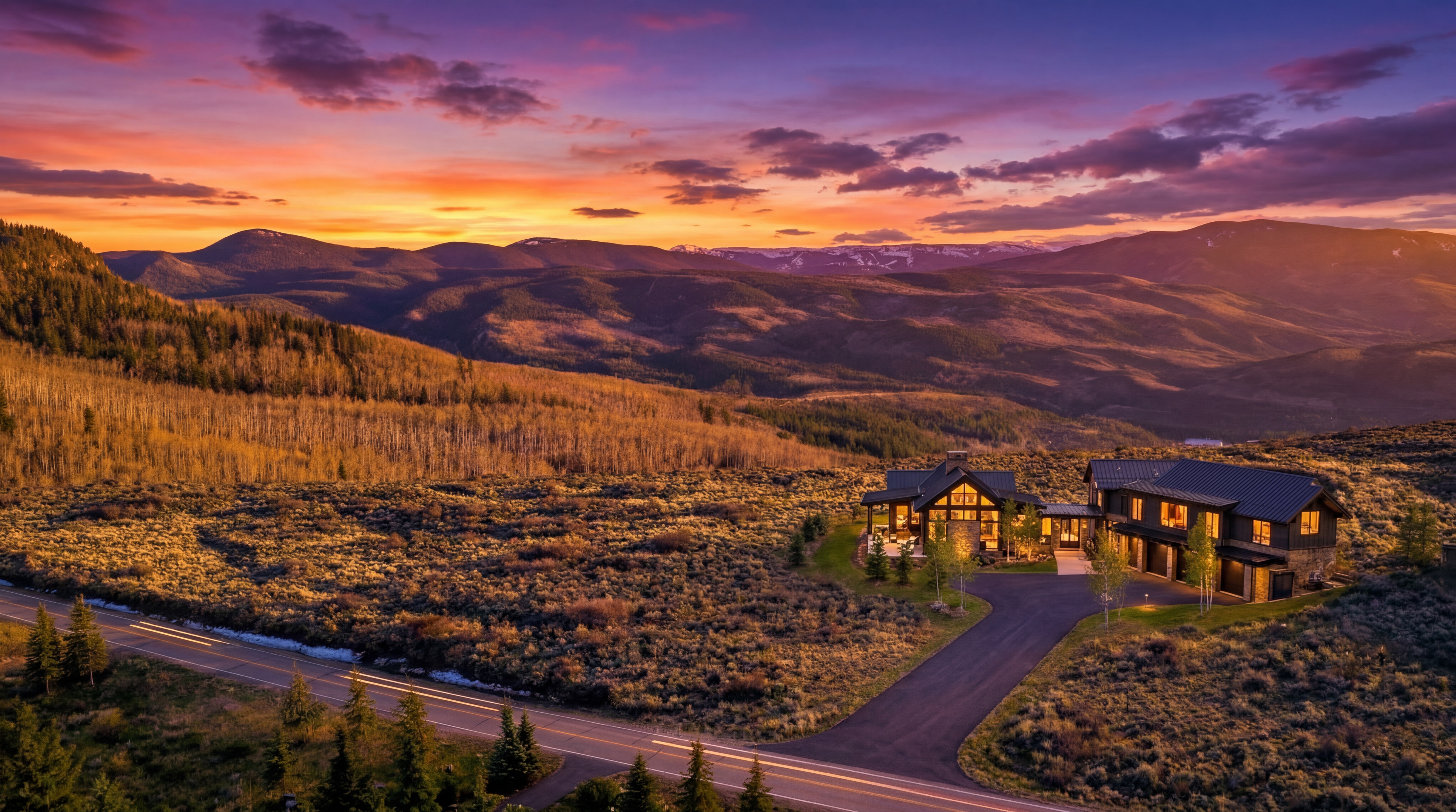 This breathtaking aerial shot captures a modern luxury mountain residence set against a vibrant, fire-hued sunset. The home features a sophisticated architectural design with dark metal roofing, warm stone accents, and expansive windows that cast a welcoming glow across the rugged landscape. The property is conveniently accessed by a paved driveway from a nearby road and is surrounded by vast, undulating mountain vistas.