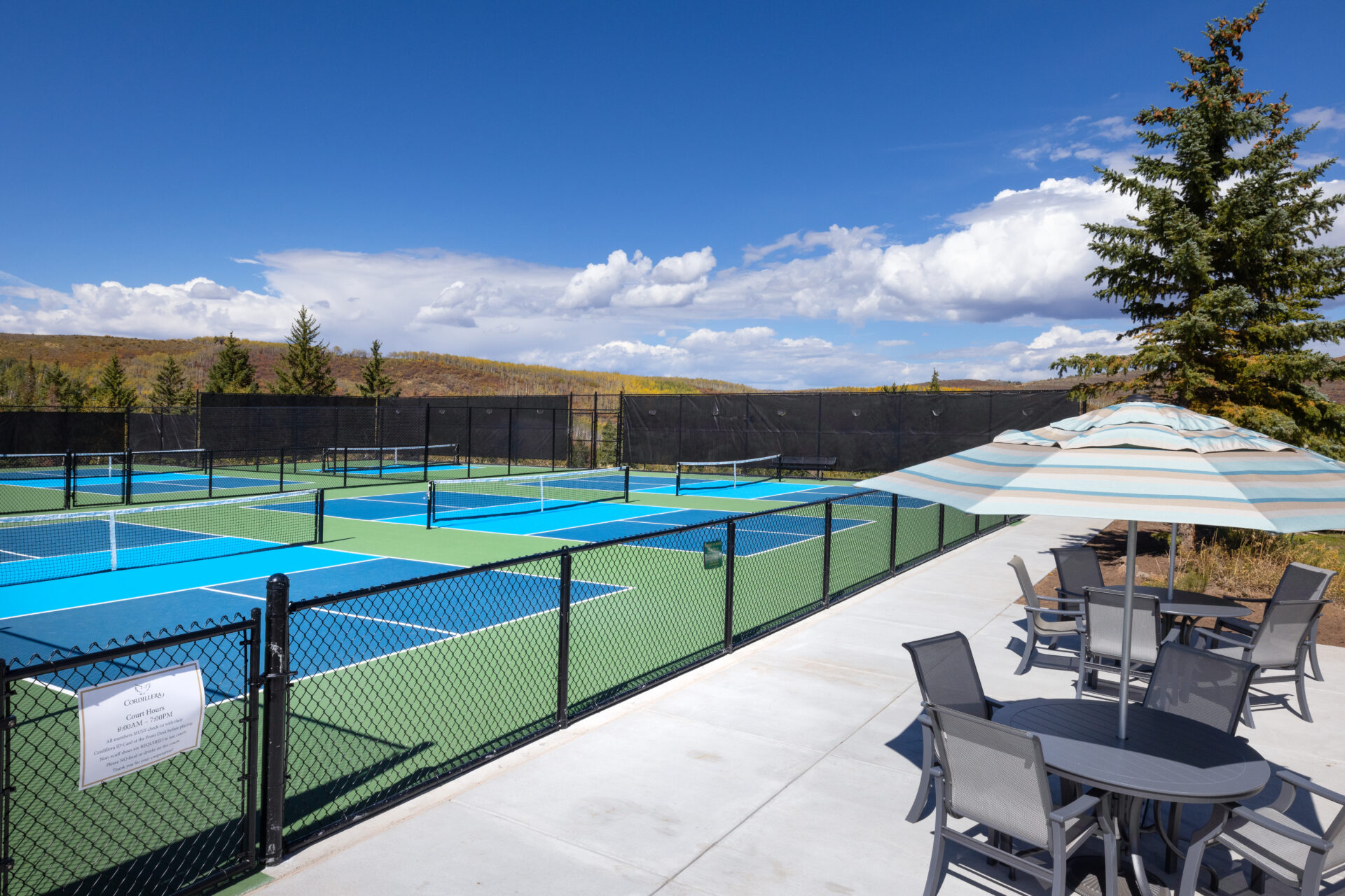 This image showcases a well-maintained community pickleball court facility set against a backdrop of scenic rolling hills under a vibrant blue sky. In the foreground, a comfortable patio area with shaded seating and outdoor furniture offers a perfect place to relax after a match. The facility is fully fenced and presents a clean, inviting space ideal for active outdoor recreation.