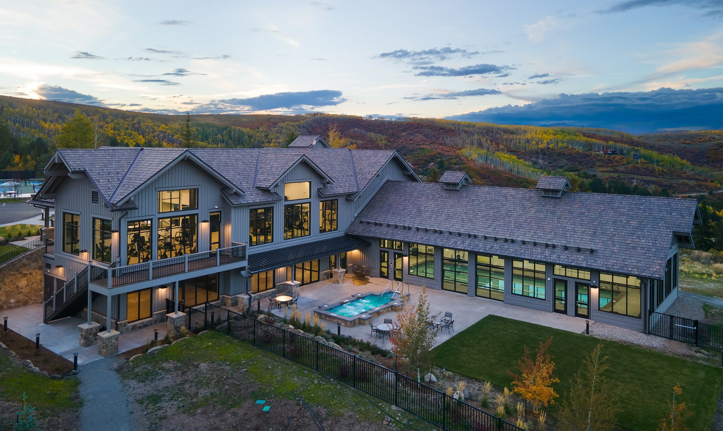 This stunning aerial view captures a large, modern clubhouse or residential common area featuring a rustic-contemporary design with grey vertical siding and dark shingle roofing. The property includes a central outdoor terrace with a built-in spa, comfortable seating, and an adjacent indoor swimming pool visible through expansive glass walls, all set against a beautiful mountain backdrop.
