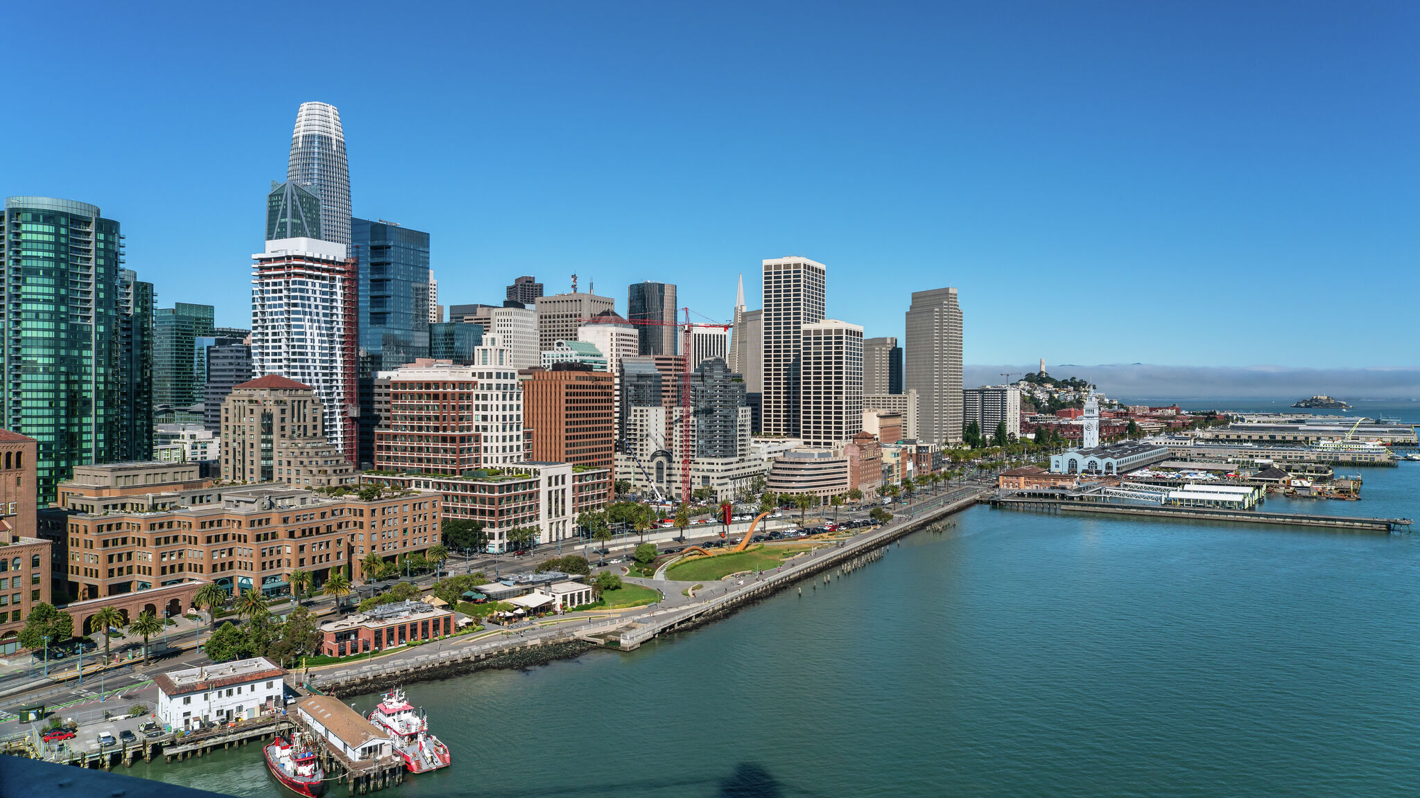 This stunning wide-angle aerial shot captures the vibrant San Francisco cityscape, showcasing a mix of modern high-rise architecture and historical waterfront buildings. The scene highlights proximity to the bay, public park spaces, and prominent local landmarks, embodying a bustling urban lifestyle. The crystal-clear blue sky and bright daylight emphasize the prominent location and expansive views offered by this metropolitan area.