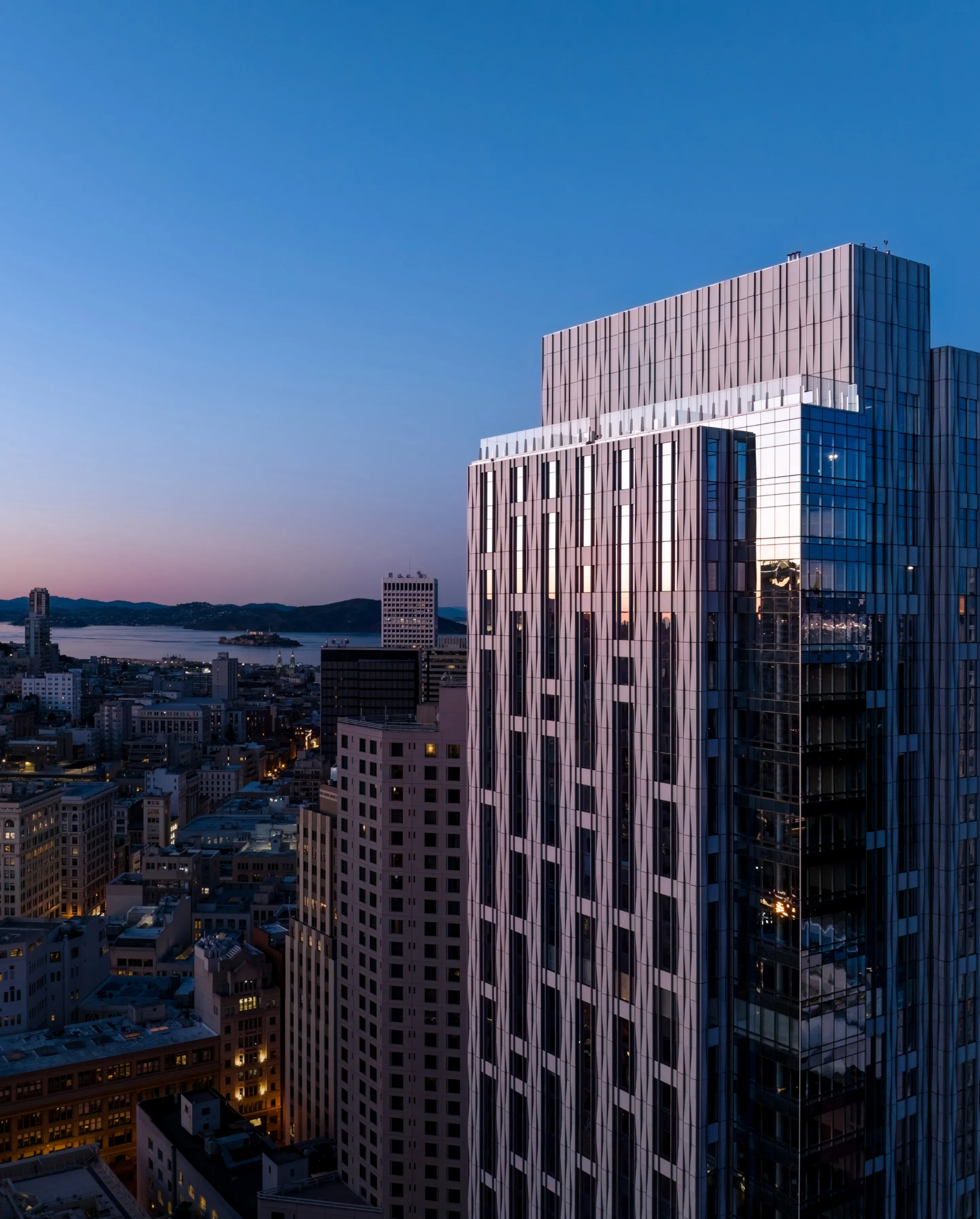 This stunning high-angle shot captures a modern skyscraper towering over the San Francisco skyline at dusk. The building features a contemporary glass and metal facade that reflects the soft twilight hues, while the surrounding urban landscape includes iconic landmarks and bay views in the distance. The high vantage point emphasizes the prime location and sophisticated architectural profile of the property.