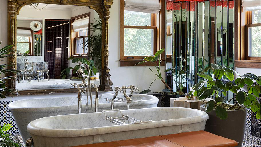 This is a luxurious primary bathroom featuring two freestanding marble bathtubs with ornate silver fixtures. A large, decorative gold-framed mirror enhances the space, reflecting intricate tilework and various plants that add a touch of nature. The overall design exudes elegance and sophistication.