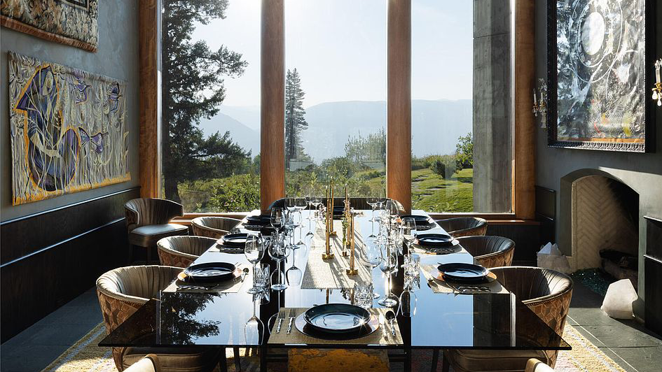 This image showcases a contemporary dining room with a large glass-topped table set for a formal meal. Floor-to-ceiling windows provide stunning views of the surrounding landscape, enhancing the dining experience. Art pieces and unique décor add to the room's sophisticated ambiance, ideal for showcasing luxury living.